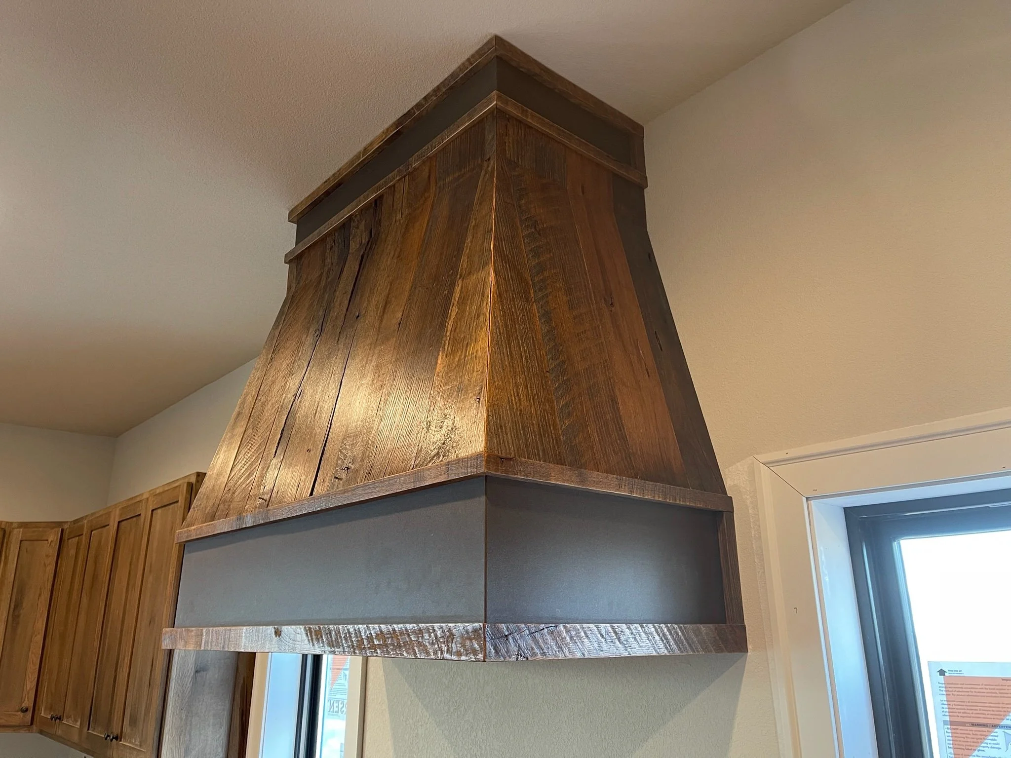 A kitchen range hood made of wood with a dark wood panel under it, mounted above a window and part of wooden kitchen cabinets.