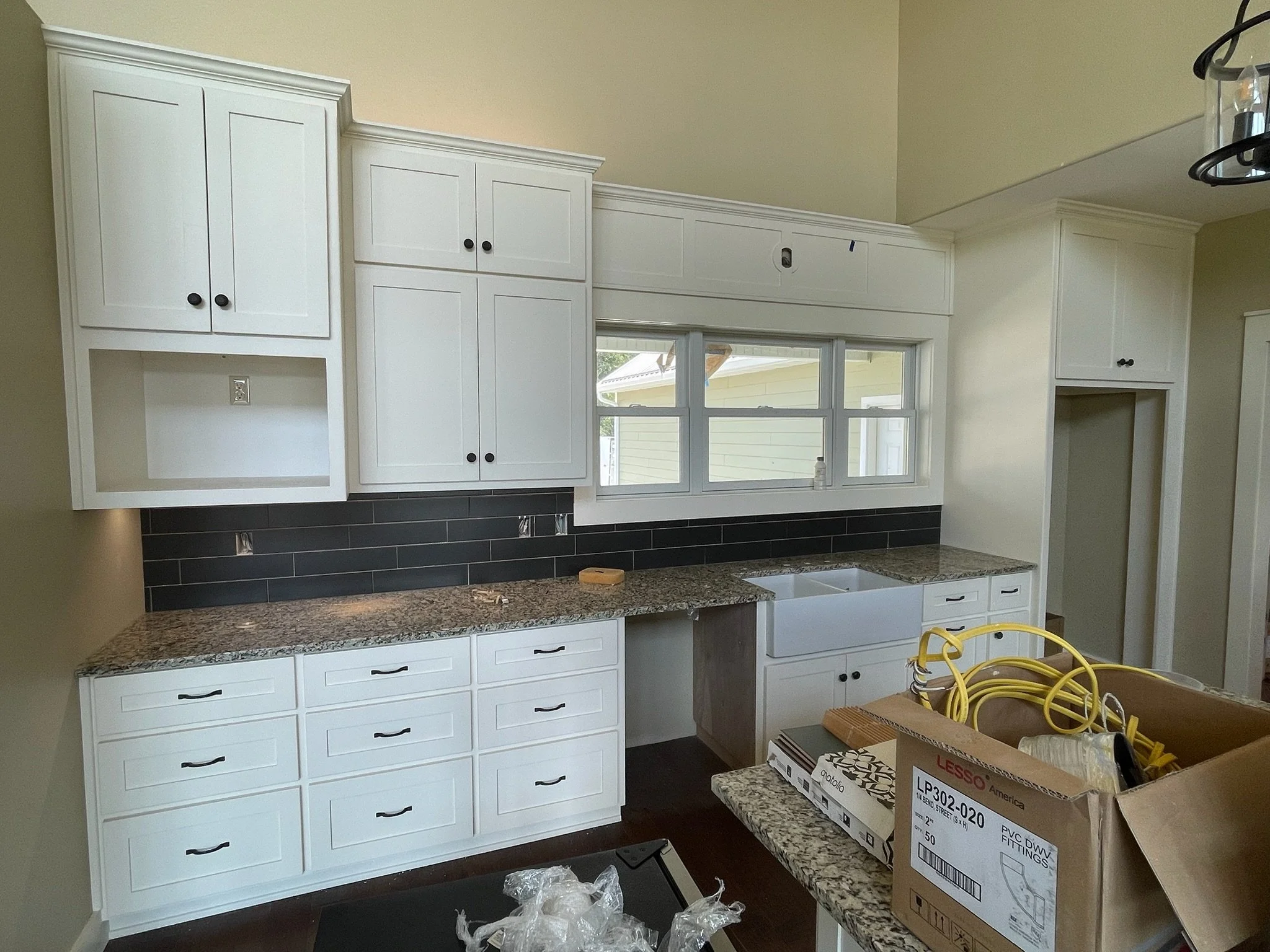 The image shows a kitchen under construction with white cabinets, granite countertops, and a black backsplash. There is a window above the sink area and some tools and materials on the counter and table.