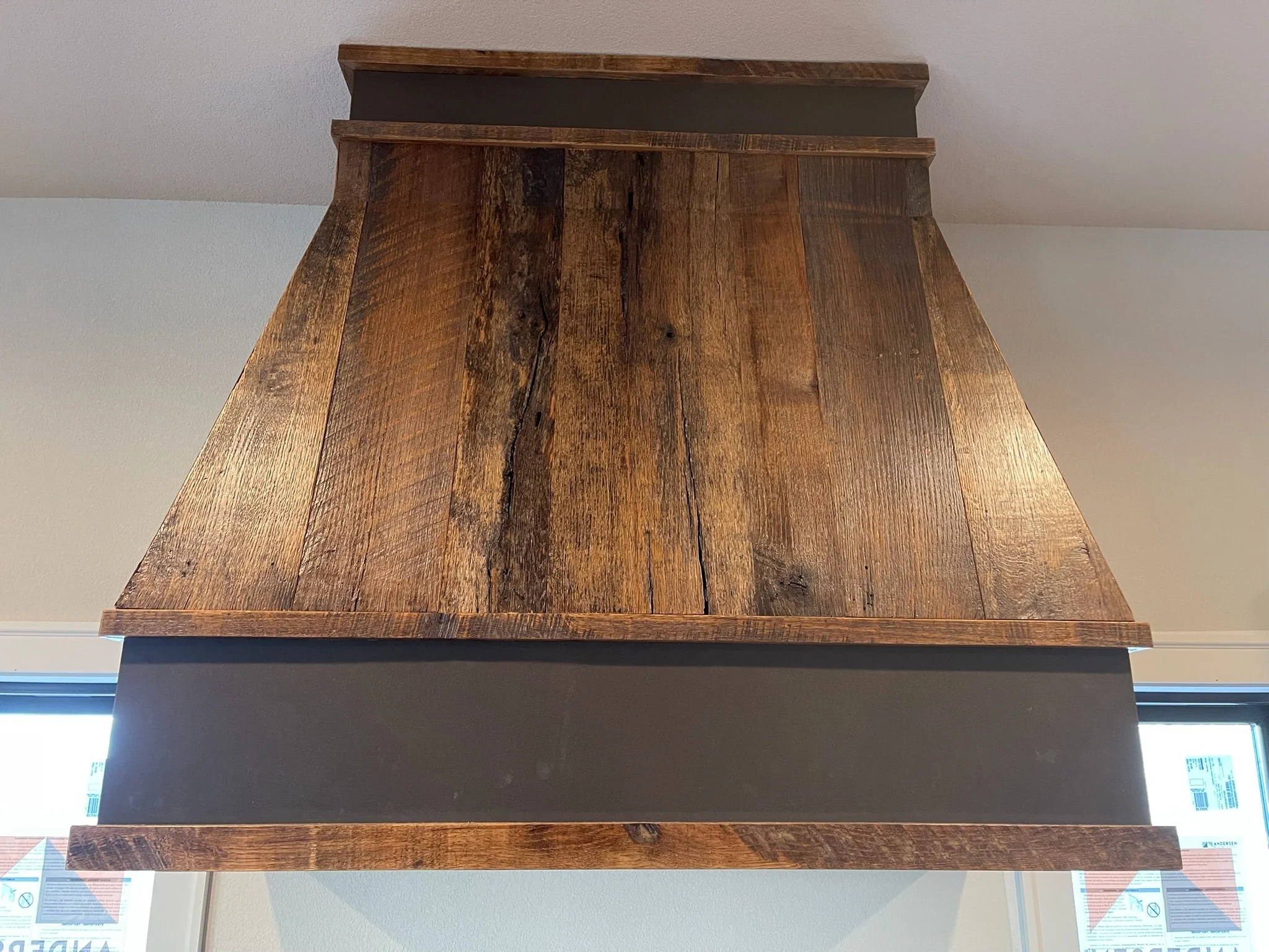 Close-up view of a wooden kitchen range hood with a dark gray panel in the center, mounted above a window with blinds.