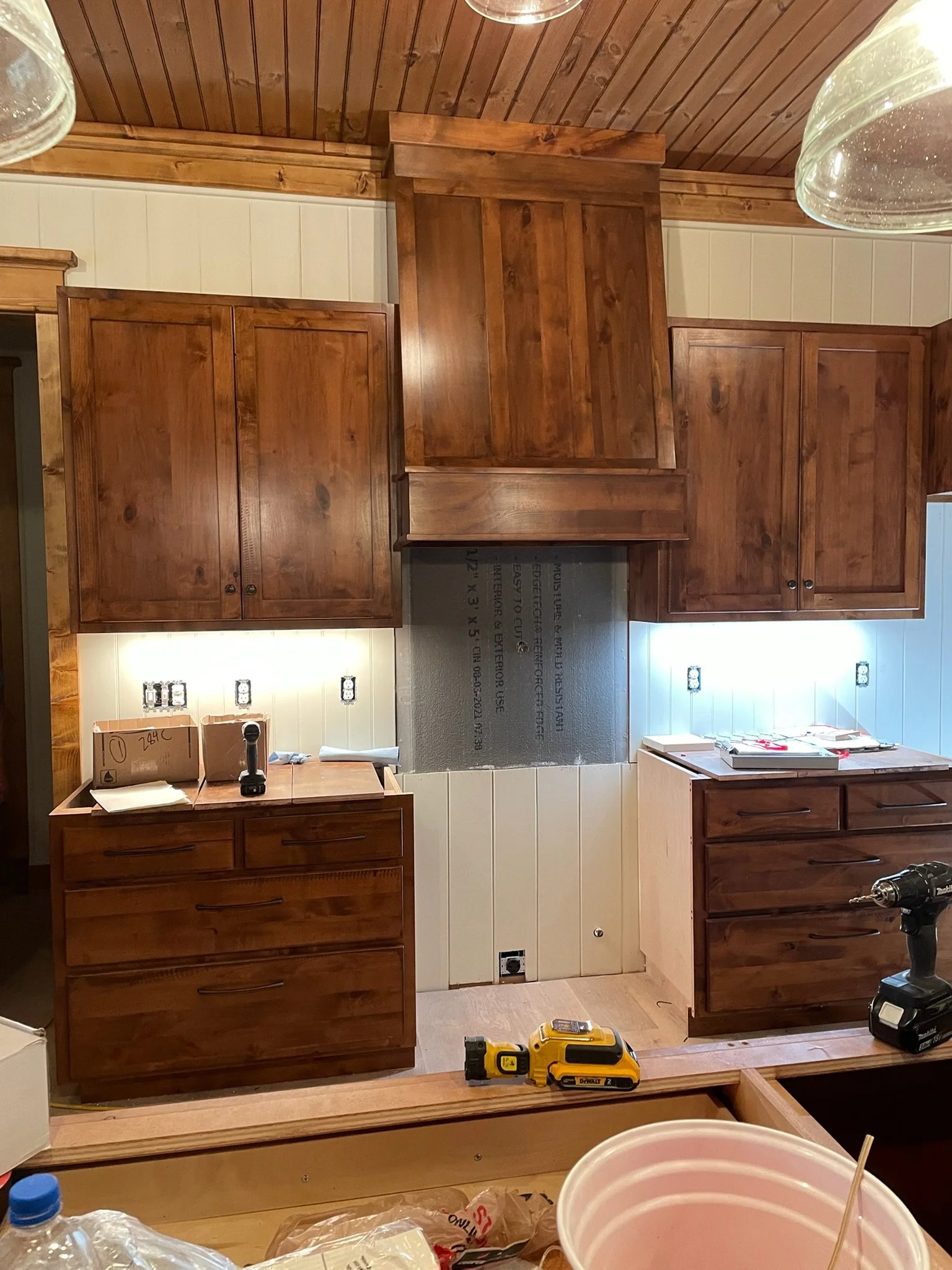 Kitchen under renovation with wooden cabinets and a range hood, electrical outlets and switches on the wall, construction tools and supplies on the countertop, and a bead of drywall visible.