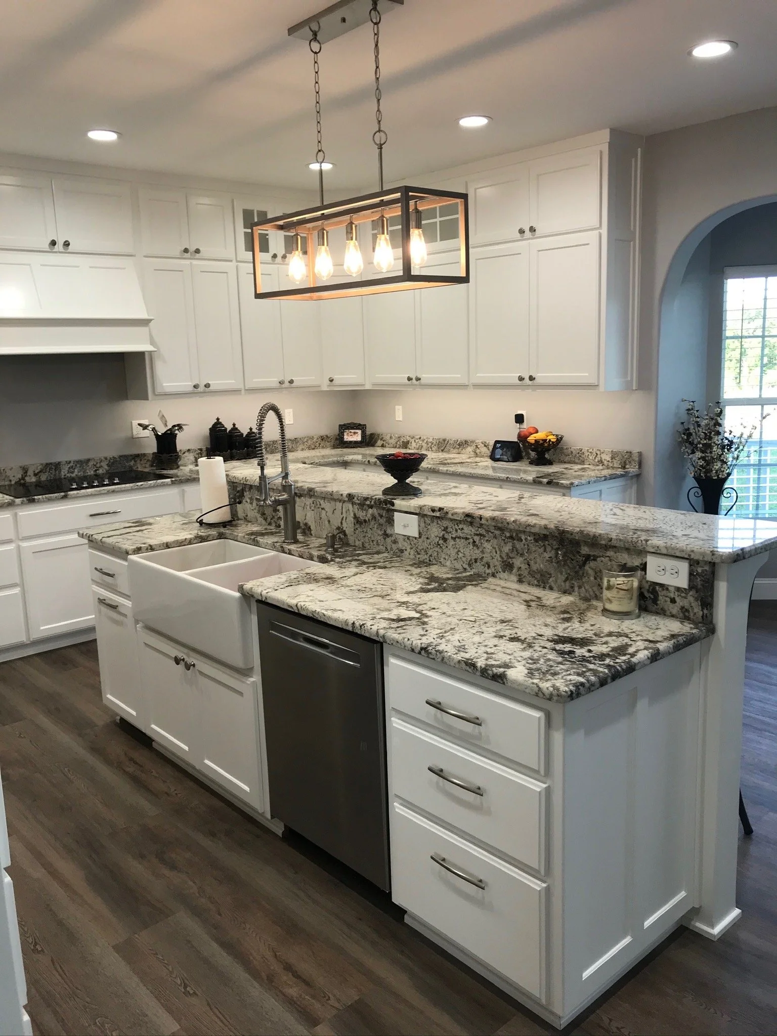 Modern kitchen with white cabinetry, granite countertops, a farmhouse sink, and a chandelier light fixture overhead. There are drawers and appliances including a small dishwasher visible under the counter. Light wood flooring and a window with sunlig