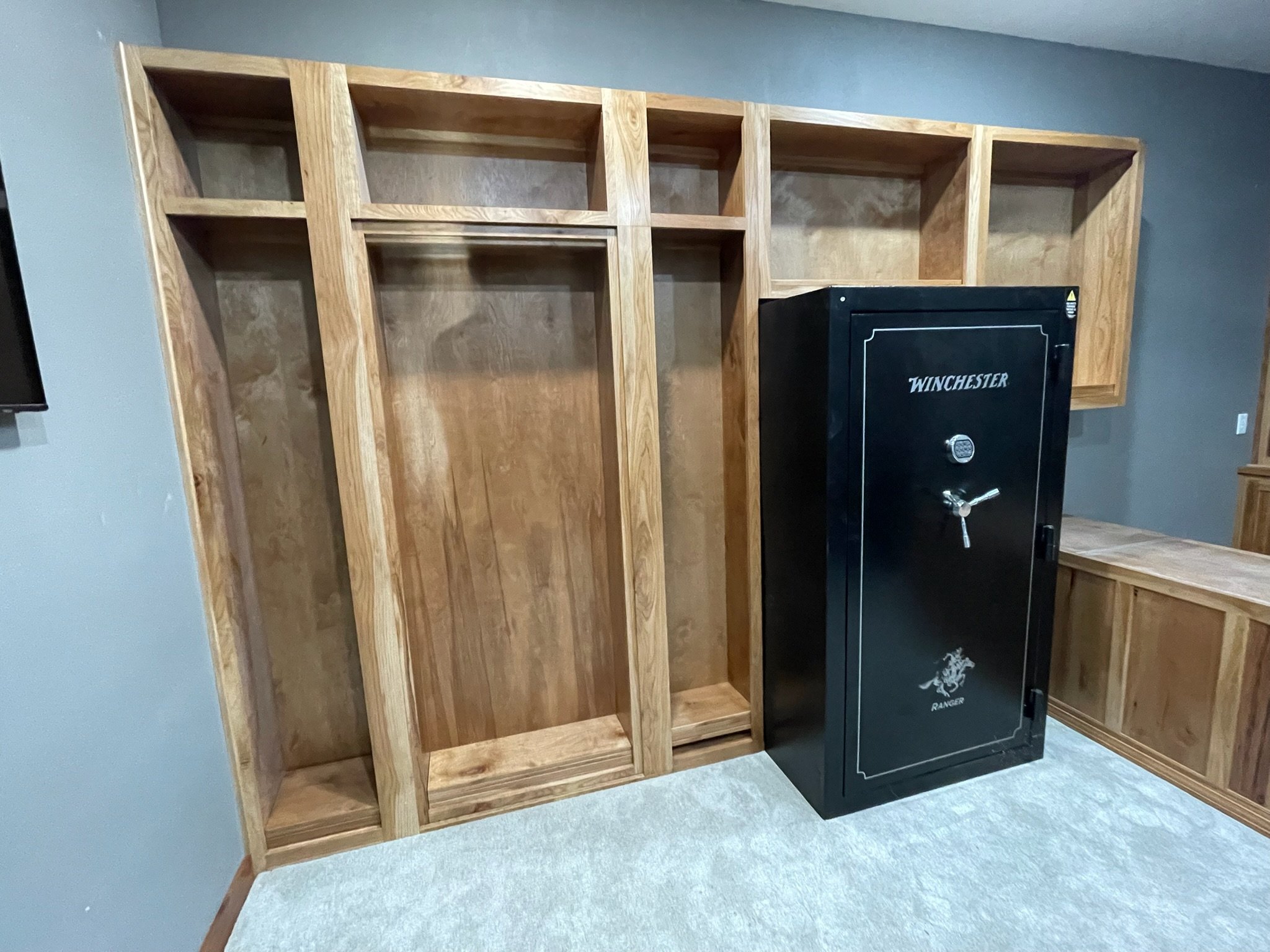 Unfinished wooden wall framing with empty shelves and a black Winchester safe in a room with carpeted floor and gray-blue walls.