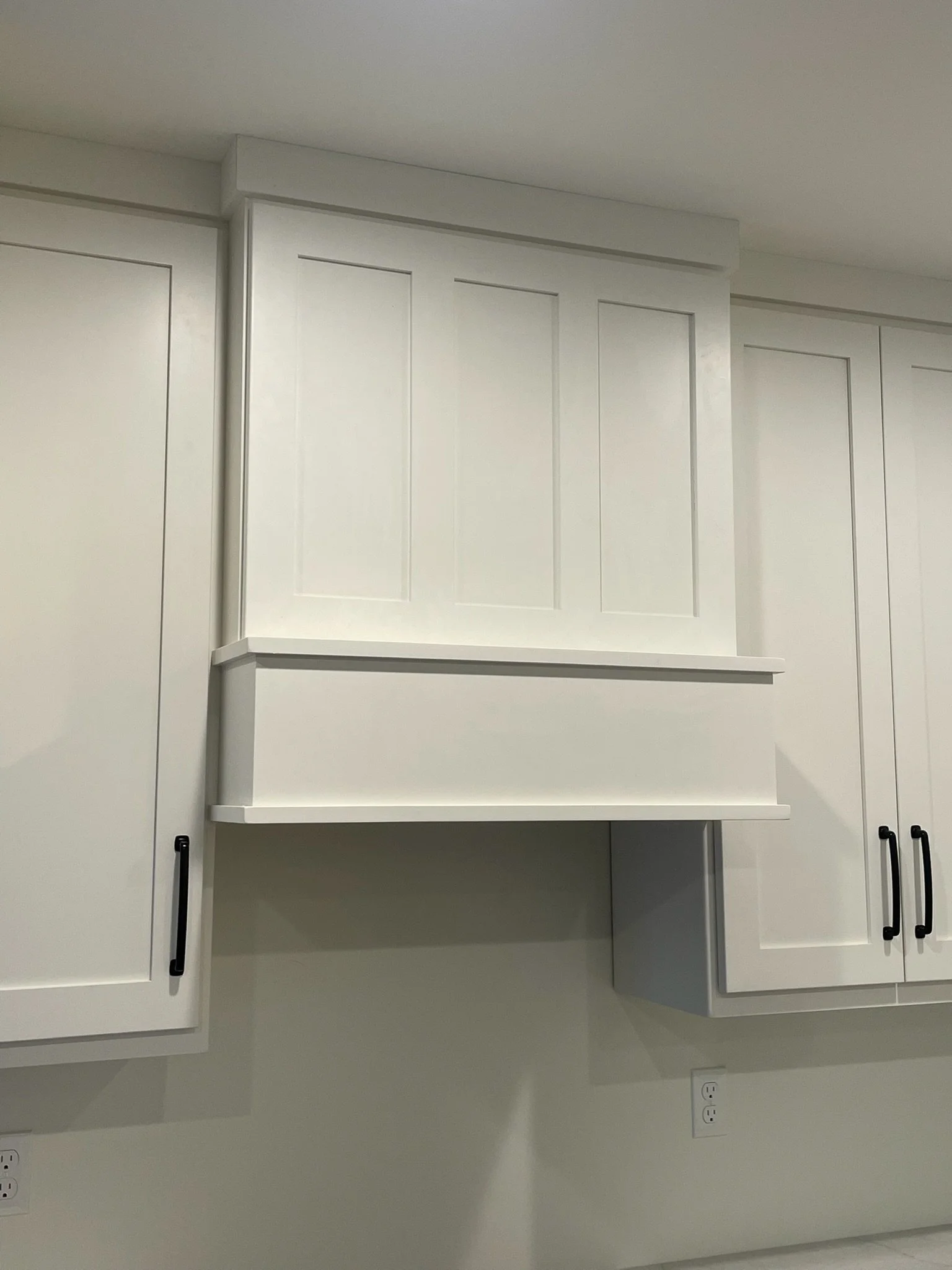 White kitchen cabinets with black handles and a small open shelf, against a plain white wall.