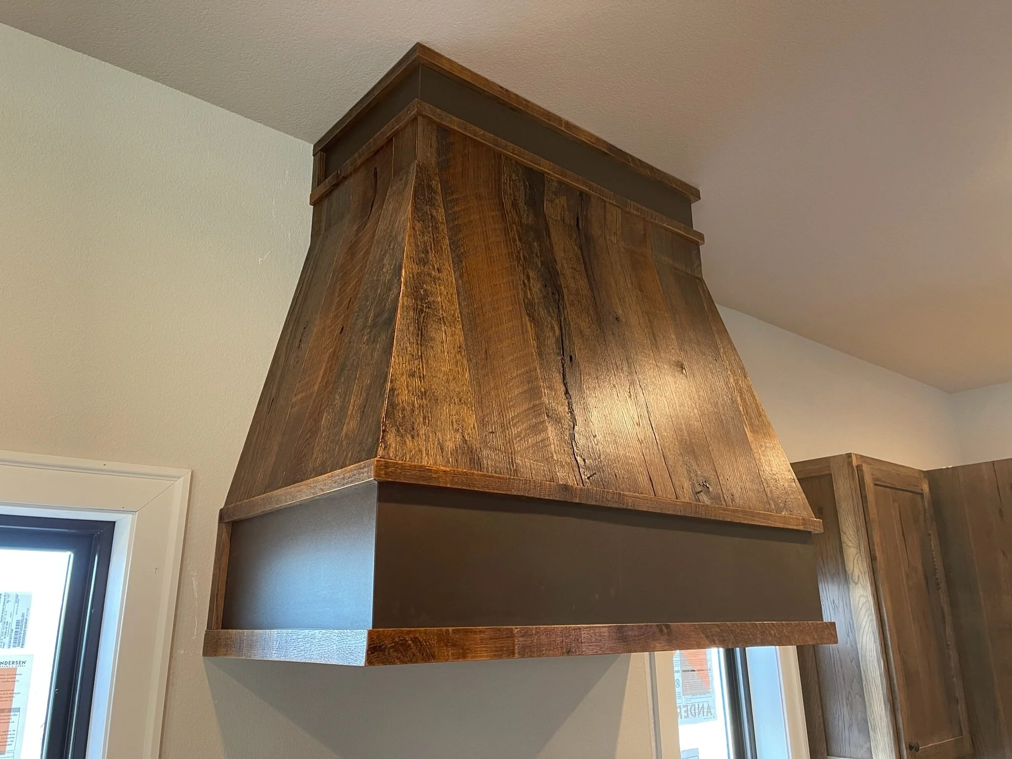 A wooden kitchen range hood with a dark metal band near the bottom, mounted above a cooking area in a kitchen with white walls and a window.