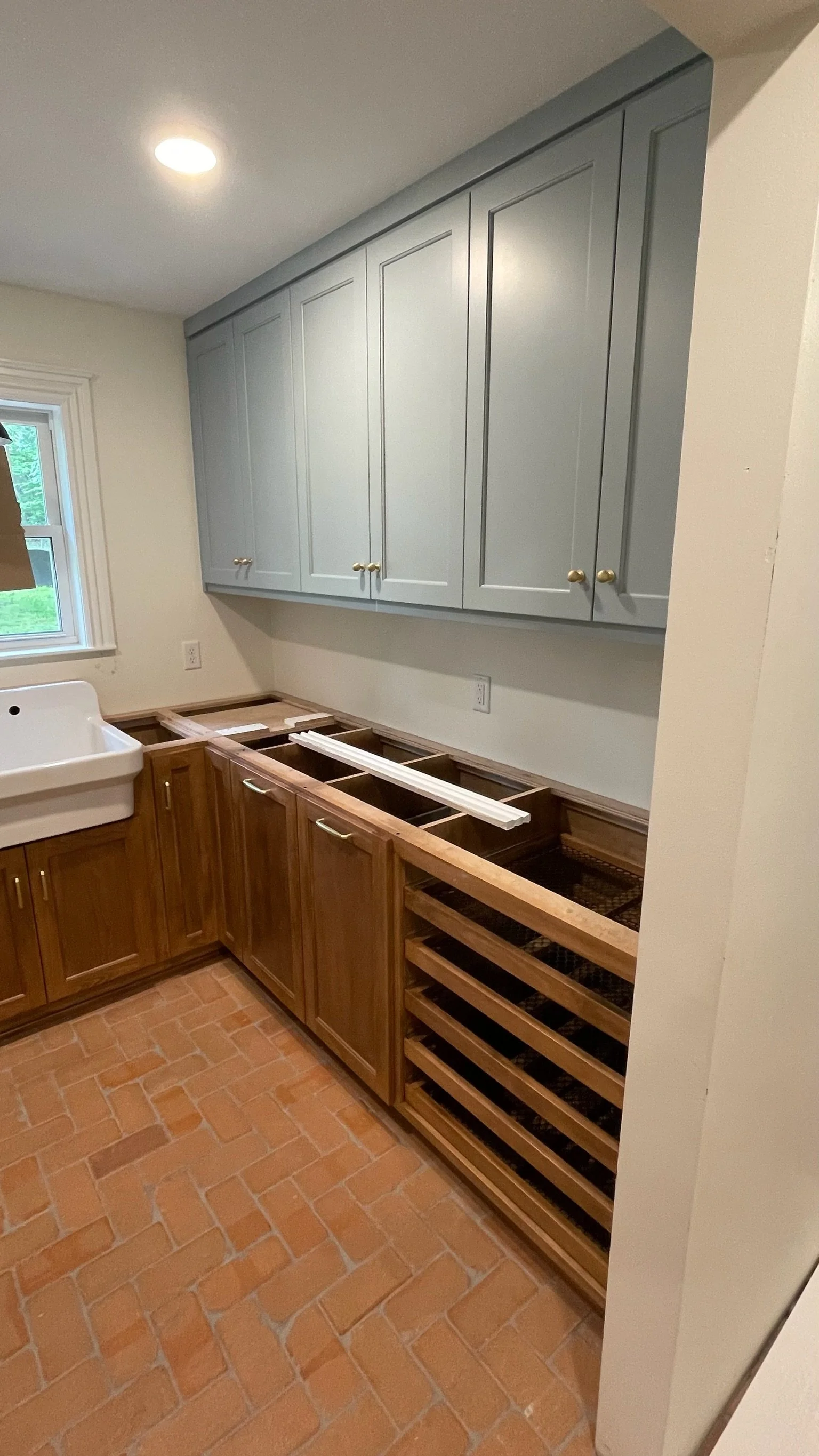 Kitchen under renovation with upper blue cabinets, lower wooden cabinets, a white farmhouse sink, and brick flooring.