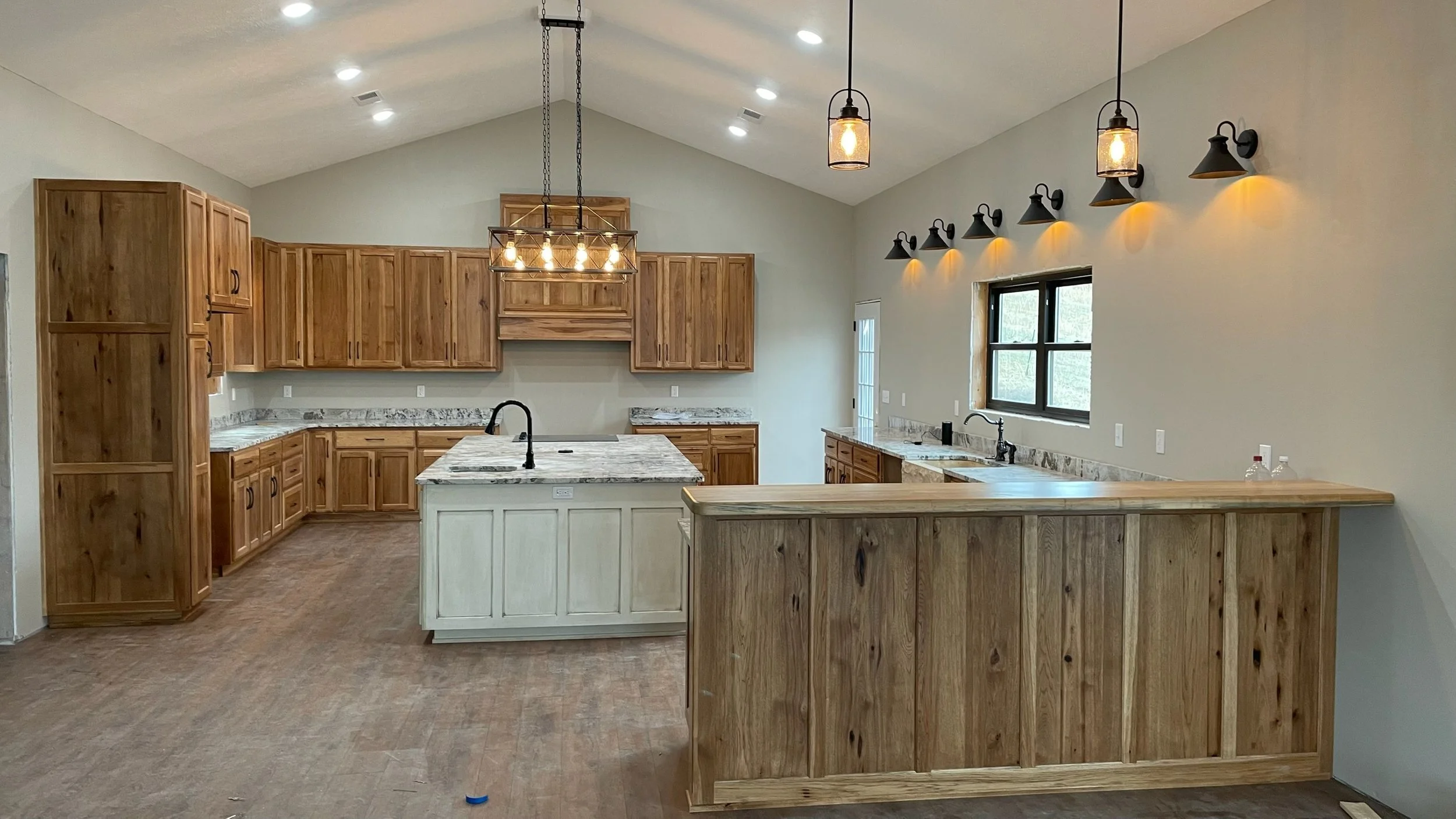 Kitchen with wooden cabinets, marble countertops, and a central island with a marble top. Overhead lighting includes a chandelier and pendant lights, with additional wall-mounted lights and a window on the right side. There are bottles on the counter