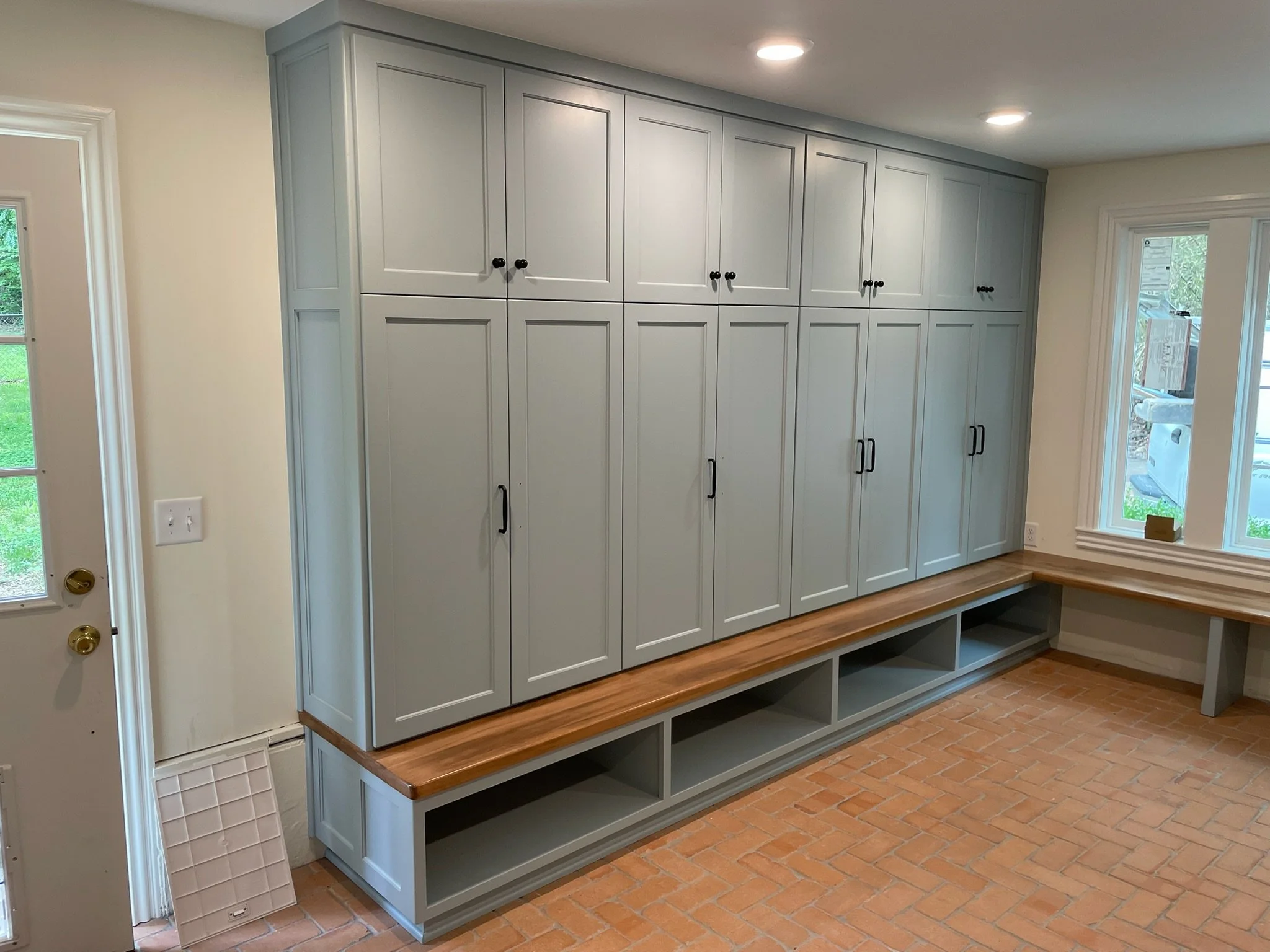 A built-in grey storage cabinet with multiple doors, black handles, and a wooden countertop, next to a window with a view outside, in a room with brick flooring.