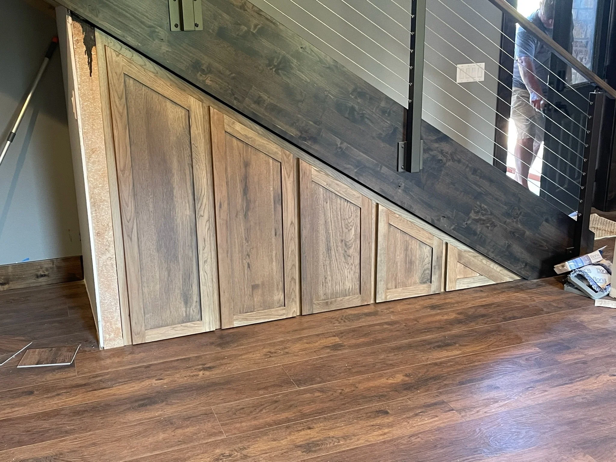 Interior view showing a staircase with wooden paneling and a metal railing, and hardwood flooring. A person is visible at the top of the stairs, partially reflected in a mirror.