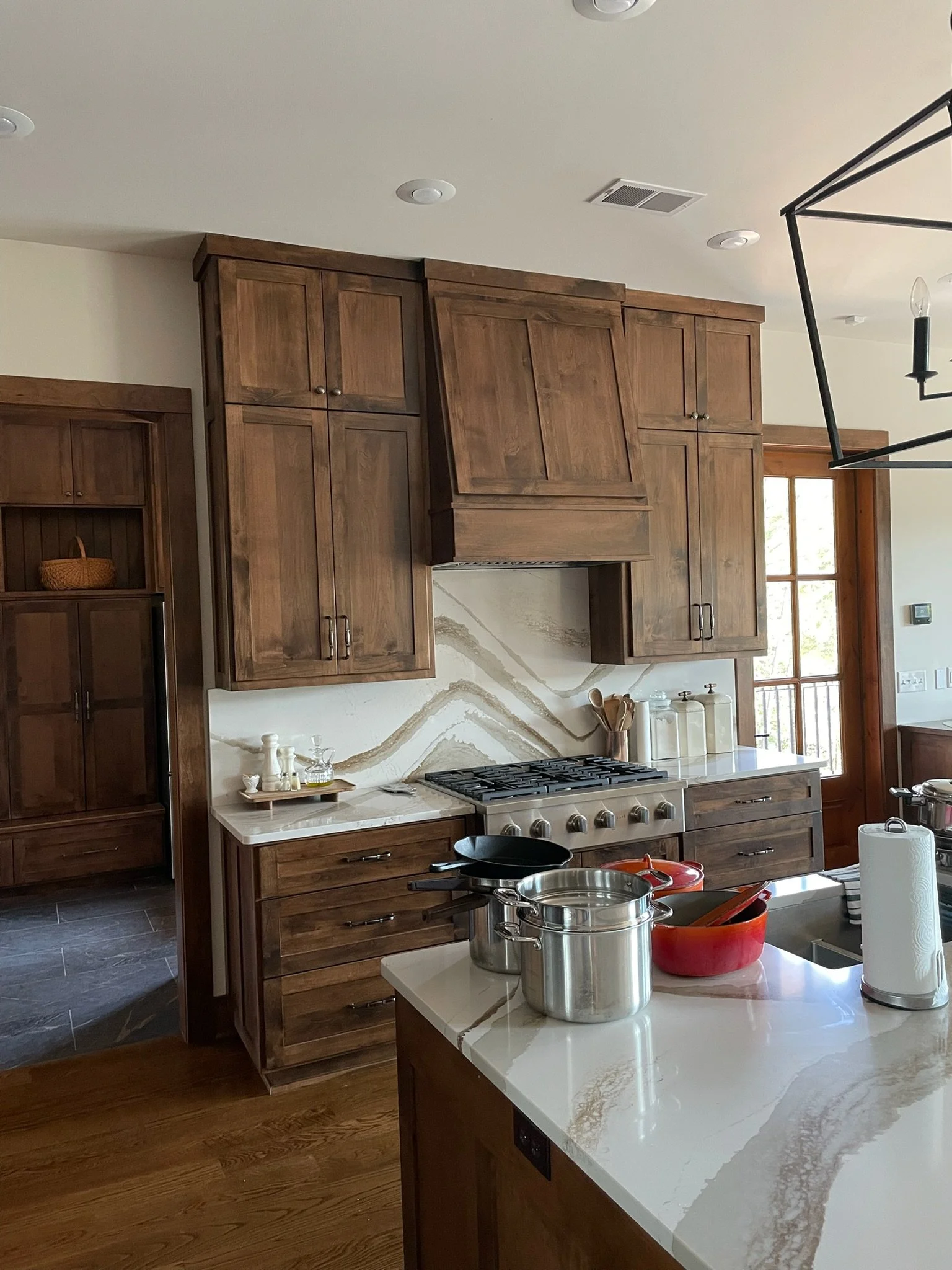 A kitchen with wooden cabinets, a marble countertop, and a stove. There are pots and pans on the island, and a paper towel roll next to a paper towel holder. A window lets in natural light.