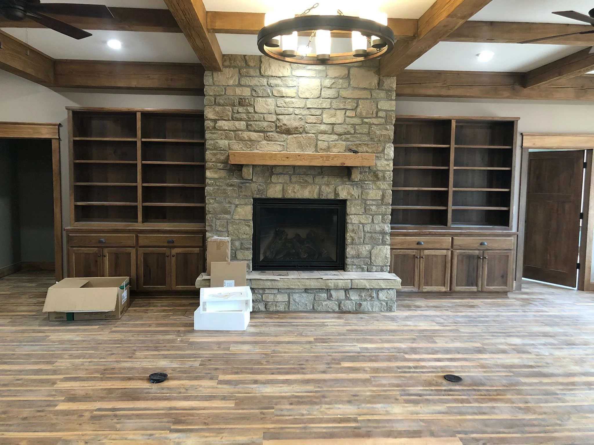 Interior room with a stone fireplace, wooden bookshelves, hardwood floor, ceiling beams, and a chandelier, with boxes on the floor.