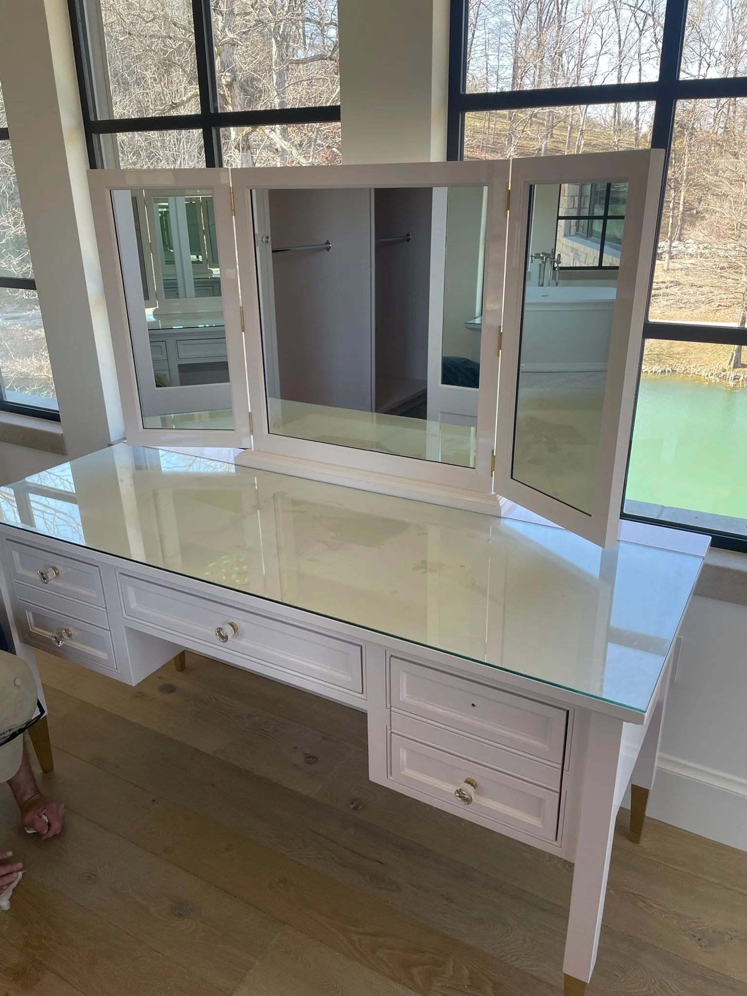 A white vanity with a large glass mirror in front of a window, showing trees outside. The vanity has several drawers with clear knobs and is situated on a hardwood floor.