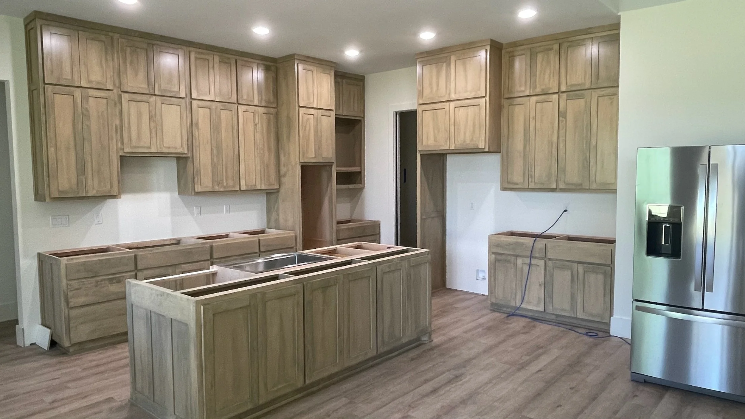 Kitchen with unfinished wooden cabinetry, a stainless steel refrigerator, and a central island with a double sink, in a room with recessed ceiling lights.