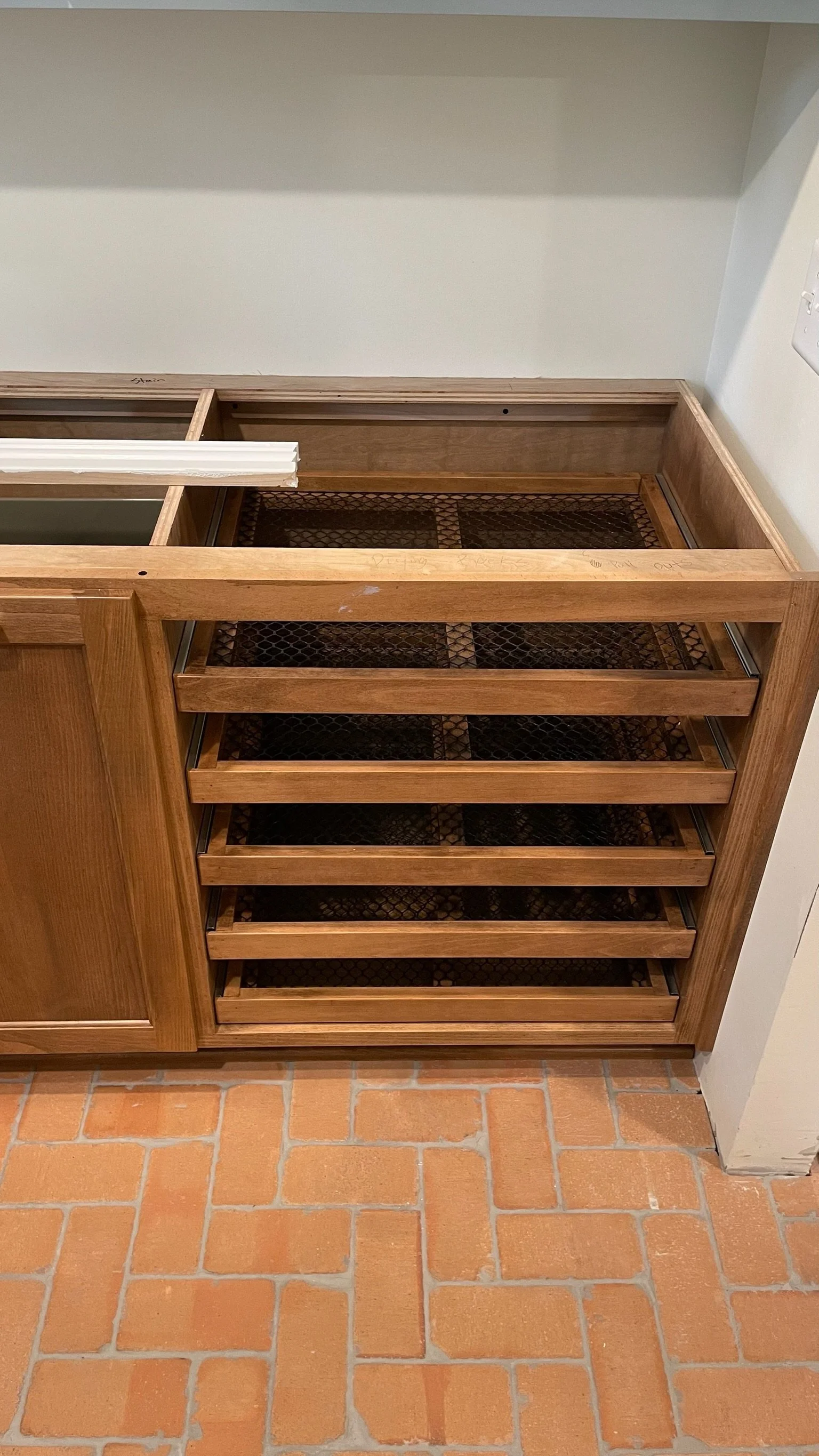 Incomplete wooden kitchen cabinet with multiple shelves and a wire mesh base, situated on a brick floor against a white wall.