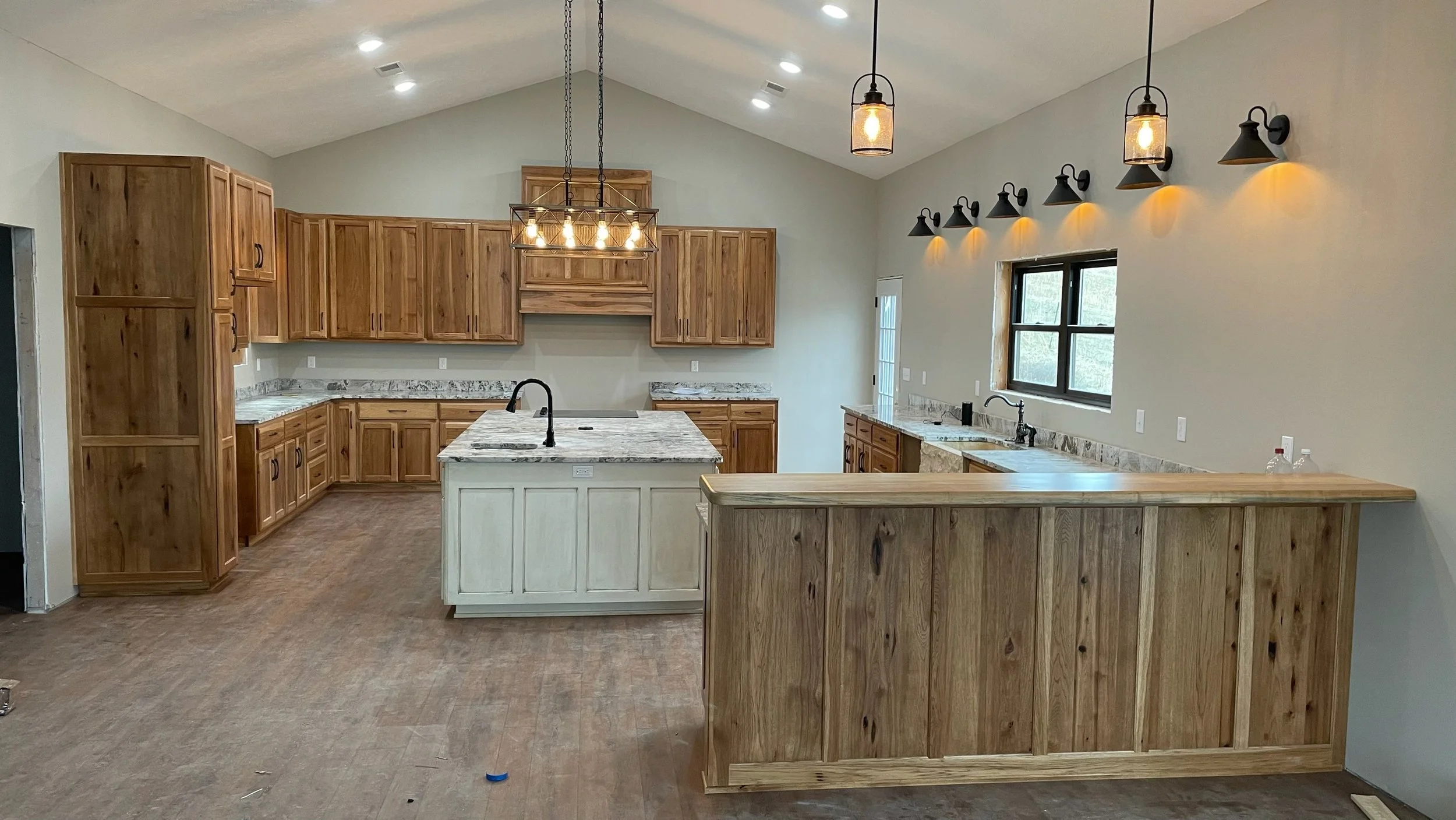 A spacious kitchen with wooden cabinets, a central island with a black faucet, granite countertops, multiple pendant and wall lights, a window above the sink, and a breakfast bar.