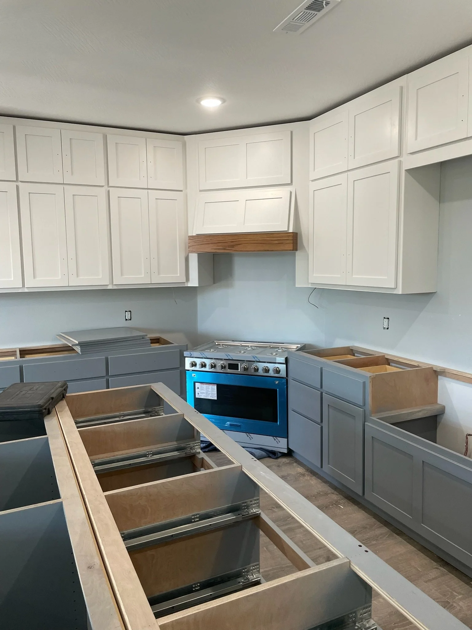 A kitchen under construction with white upper cabinets, gray lower cabinets, and a stainless steel oven. The countertop is unfinished.