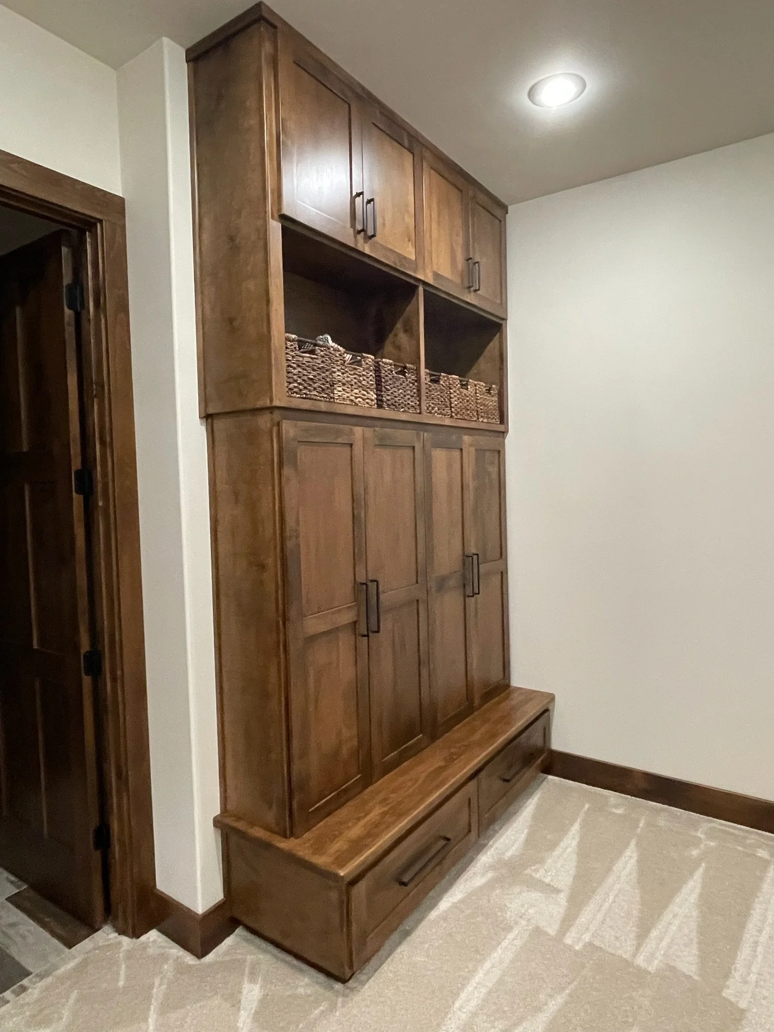 A tall wooden storage cabinet with multiple closed cabinet doors and open cubbies, situated against a white wall in a room with light-colored carpet.