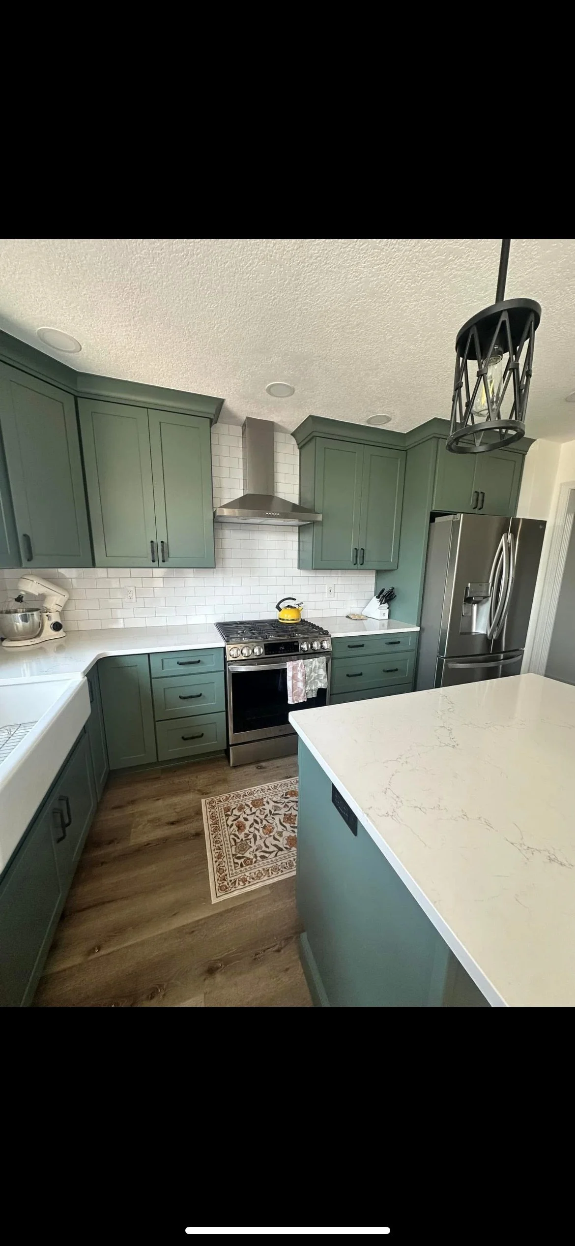 A modern kitchen with green cabinets, stainless steel appliances including a refrigerator and an oven, white subway tile backsplash, a white marble island, and a patterned rug on wood flooring. There's a black hanging light fixture and a yellow kettl