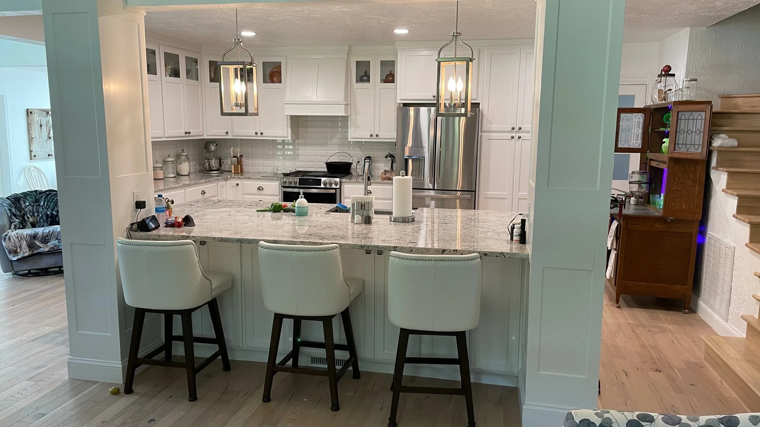 A kitchen with white cabinets, a granite island counter, and a modern stainless steel refrigerator. The island has three white chairs, and there are pendant lights hanging above. To the left, part of a living area with a floral couch is visible. To t