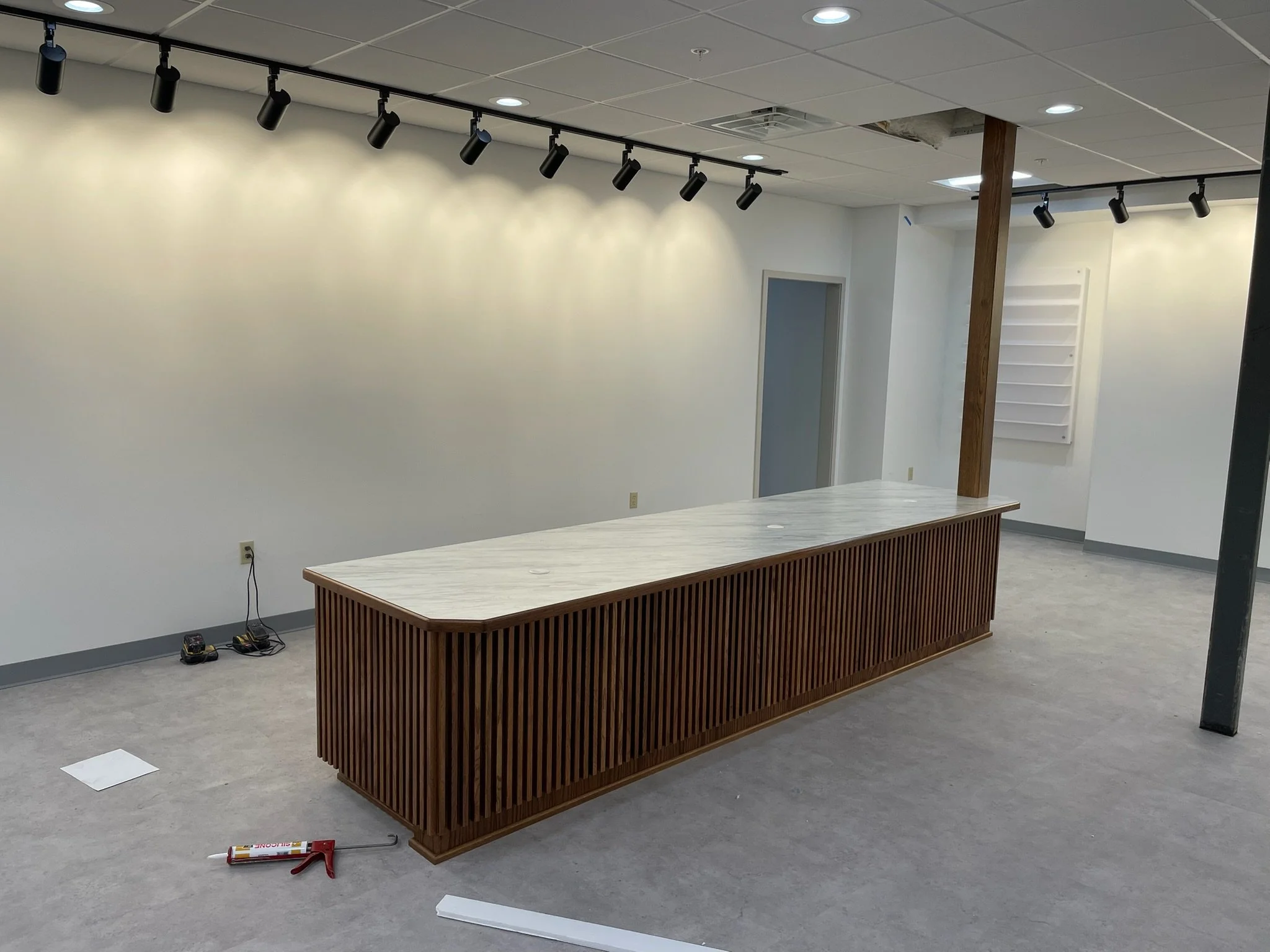 Empty room with a long wooden and marble countertop, track lighting on the ceiling, and some construction tools on the floor.