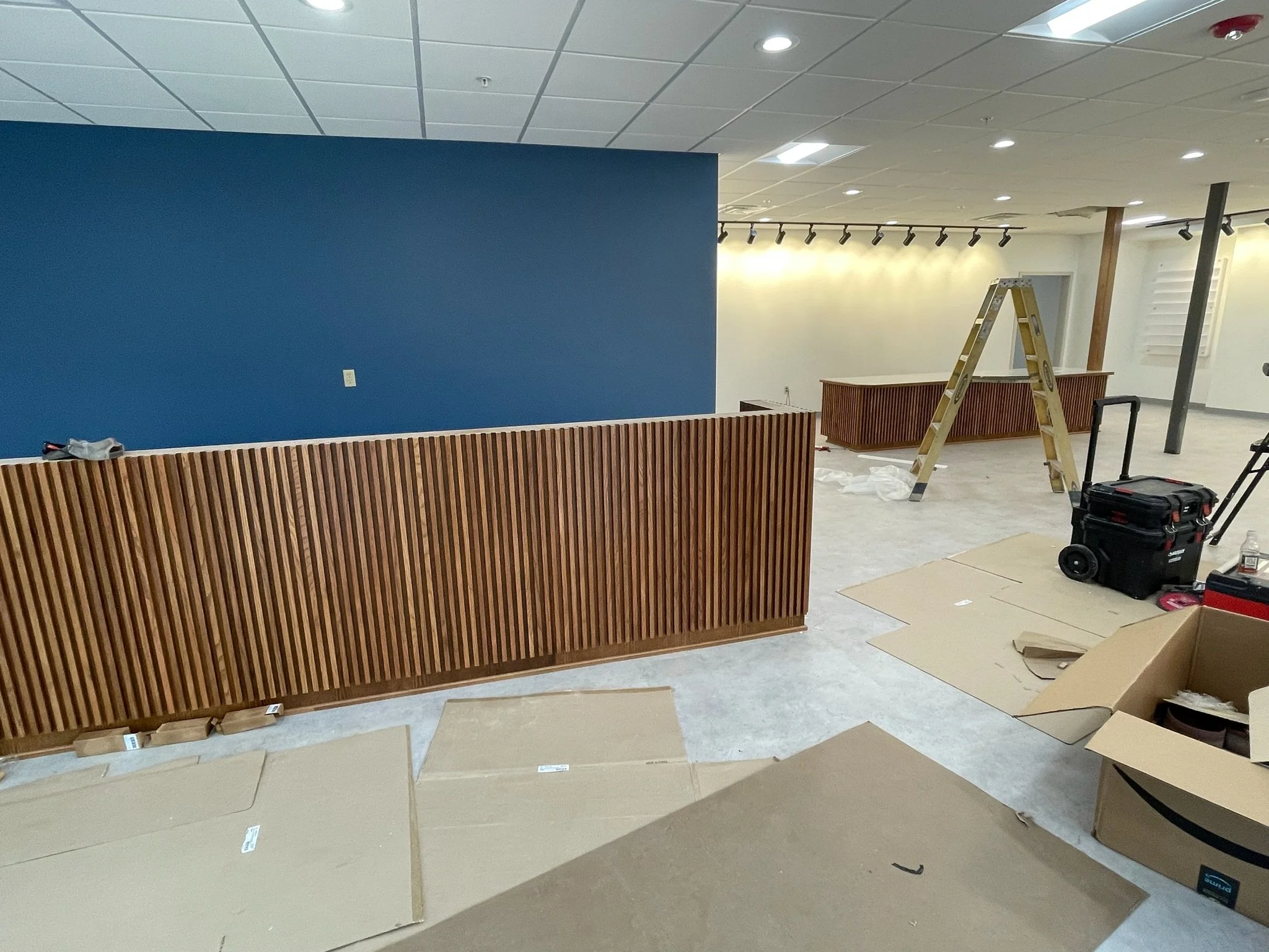 Interior of a commercial space under construction with a blue accent wall, wooden wainscoting, a ladder, construction tools, and packaging materials.