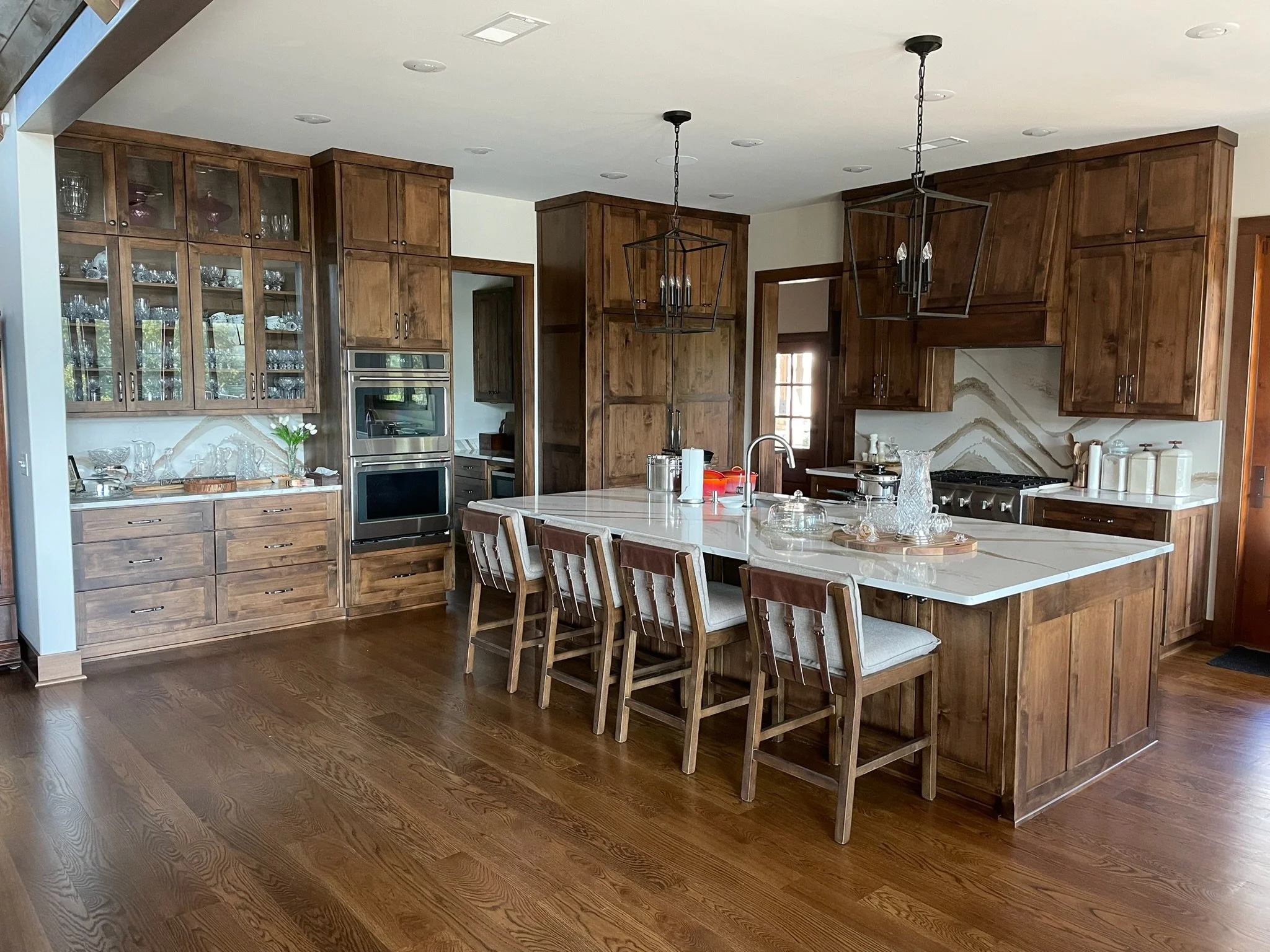 Modern kitchen with wooden cabinets, white marble island, and hardwood floors. Features built-in double ovens, glass-front upper cabinets, and pendant lighting over the island. Kitchen decor includes glass vases and white containers.