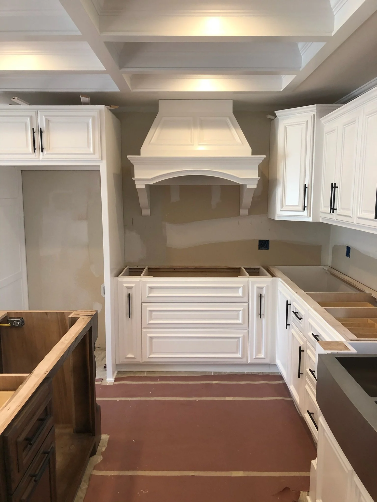 Kitchen under renovation with white cabinets, a large wooden countertop frame, and a honey-colored floor. Construction materials and tools are visible.