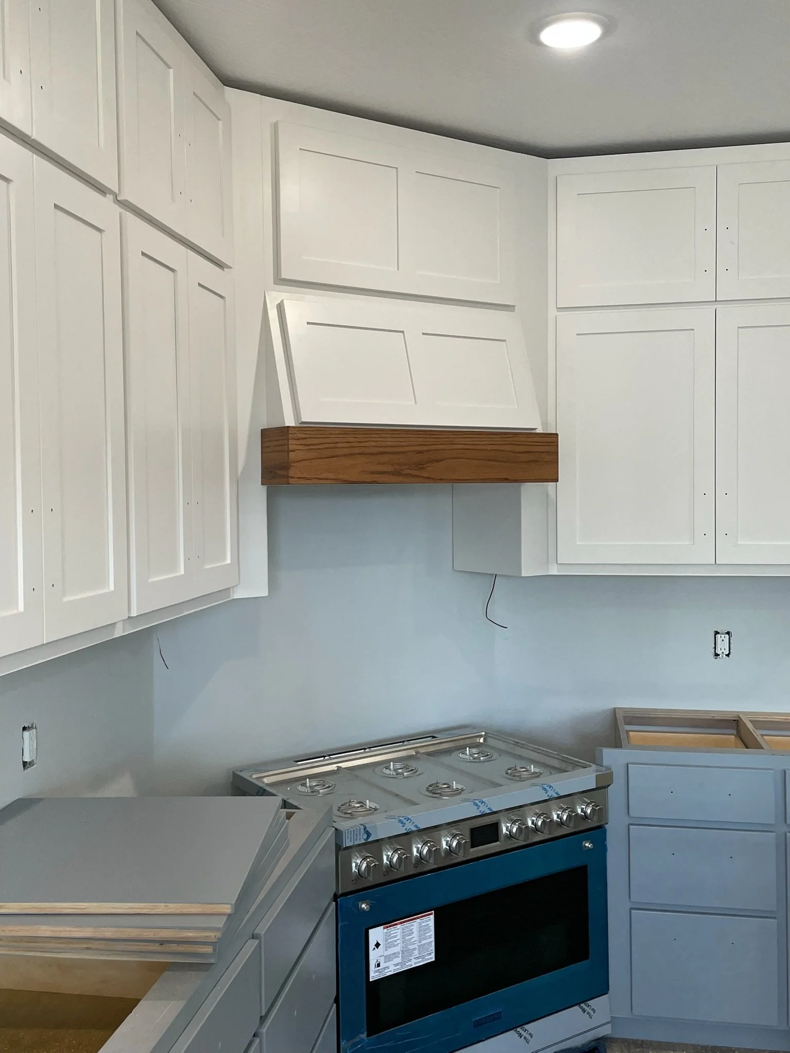 An unfinished kitchen with white cabinets, an uninstalled stove, and a vent hood. There is a partially assembled cabinet on the right and exposed wiring on the wall.