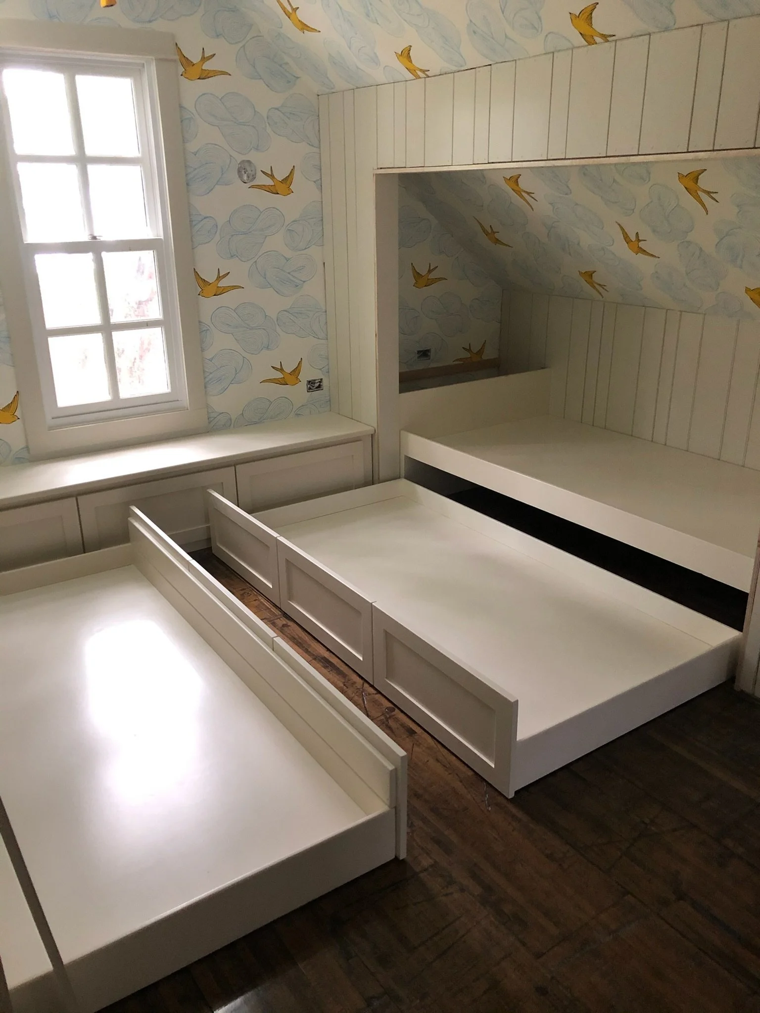 Bedroom with a partially assembled white bed frame, a window, and wall with wallpaper featuring blue clouds and flying yellow birds.