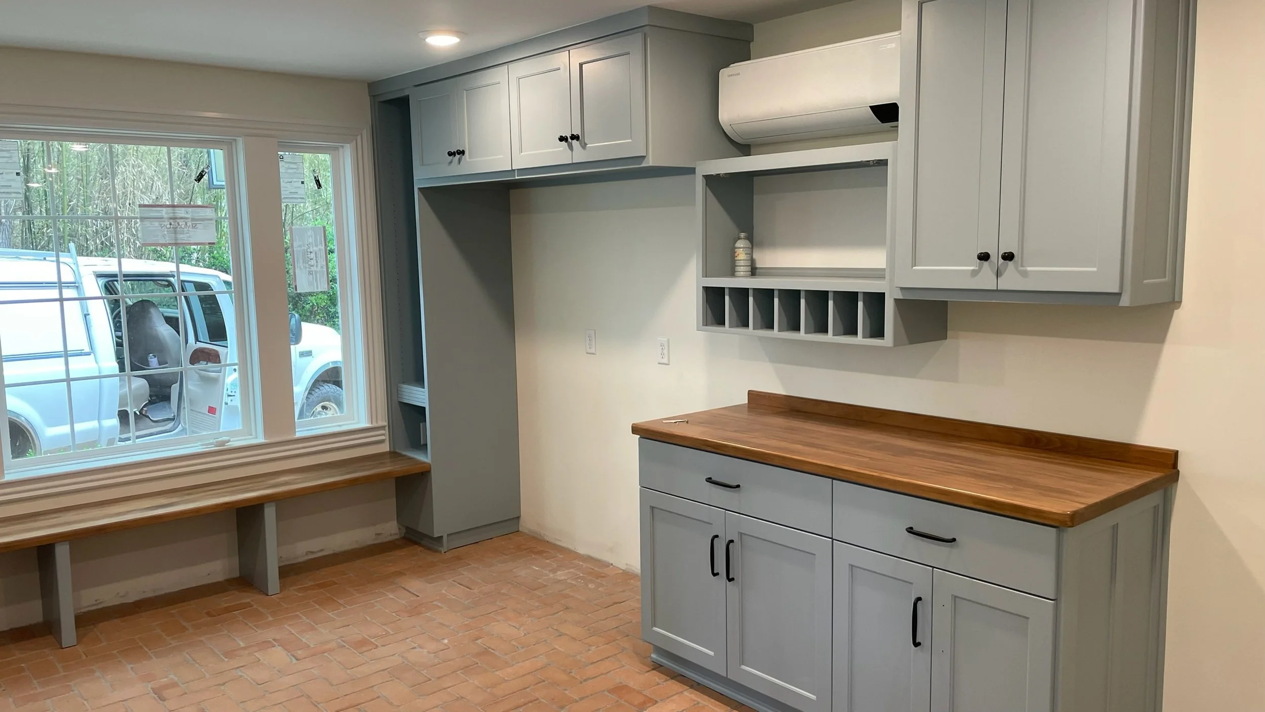 Empty kitchen area with gray cabinets, wooden countertops, brick floor, windows, and a bench by the window. Outside, white van and greenery visible.