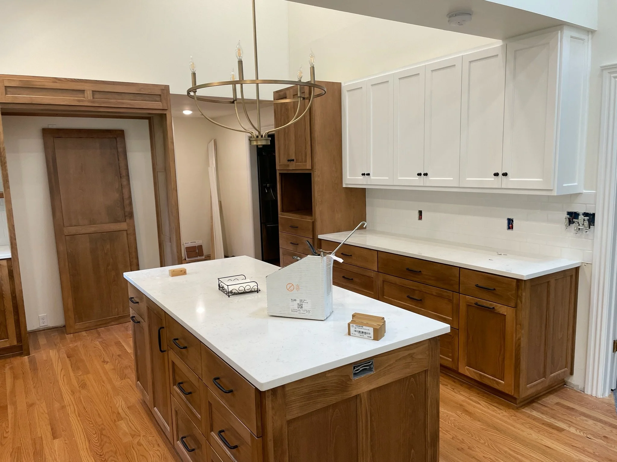 Modern kitchen with wooden cabinets, white countertops, and a central island. There is a chandelier hanging above the island, and some furniture and construction materials on the island, indicating ongoing work.
