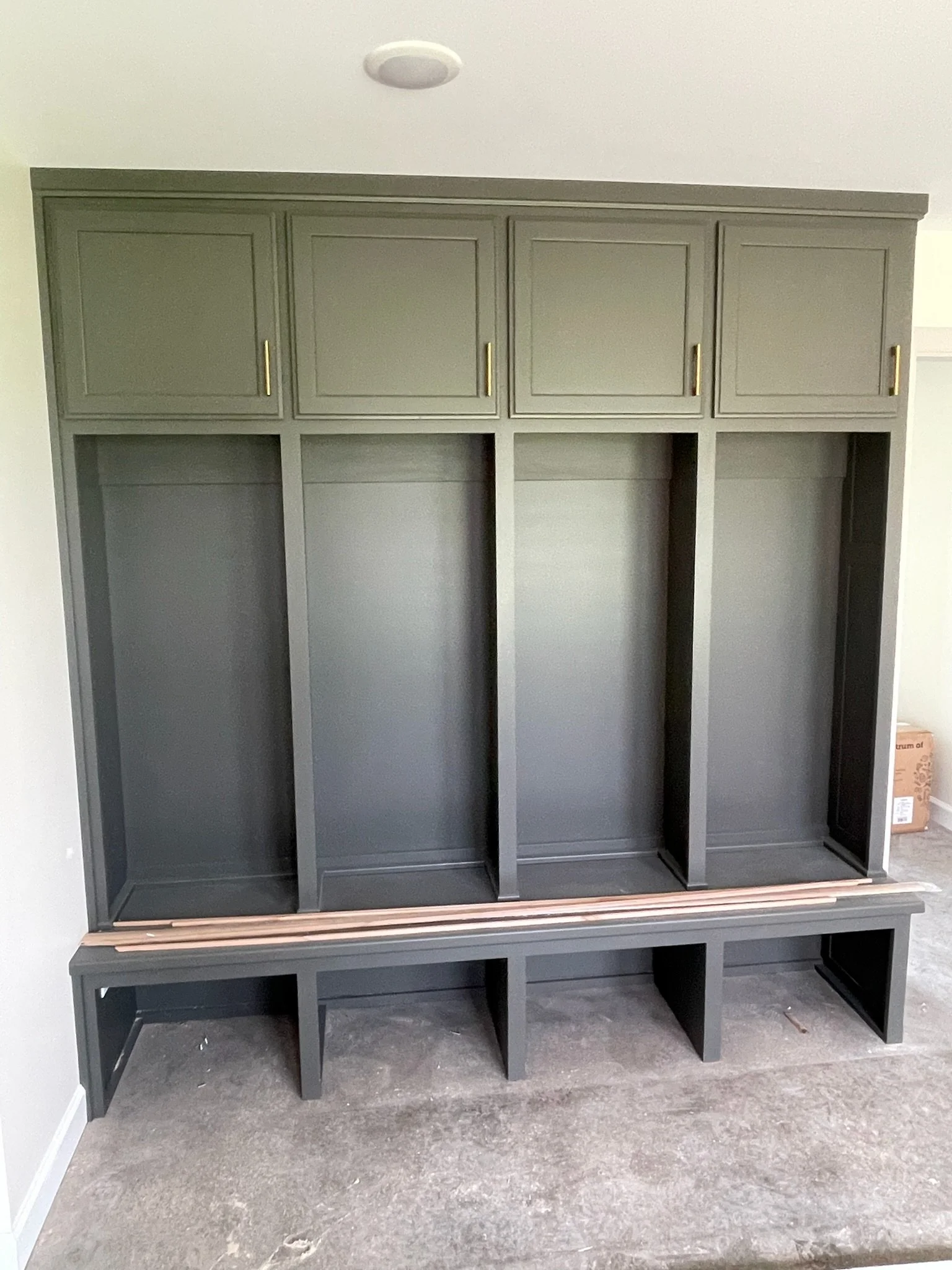 Unfinished gray built-in entryway storage with cabinets above and open cubbies below, on a concrete floor in a room under construction.