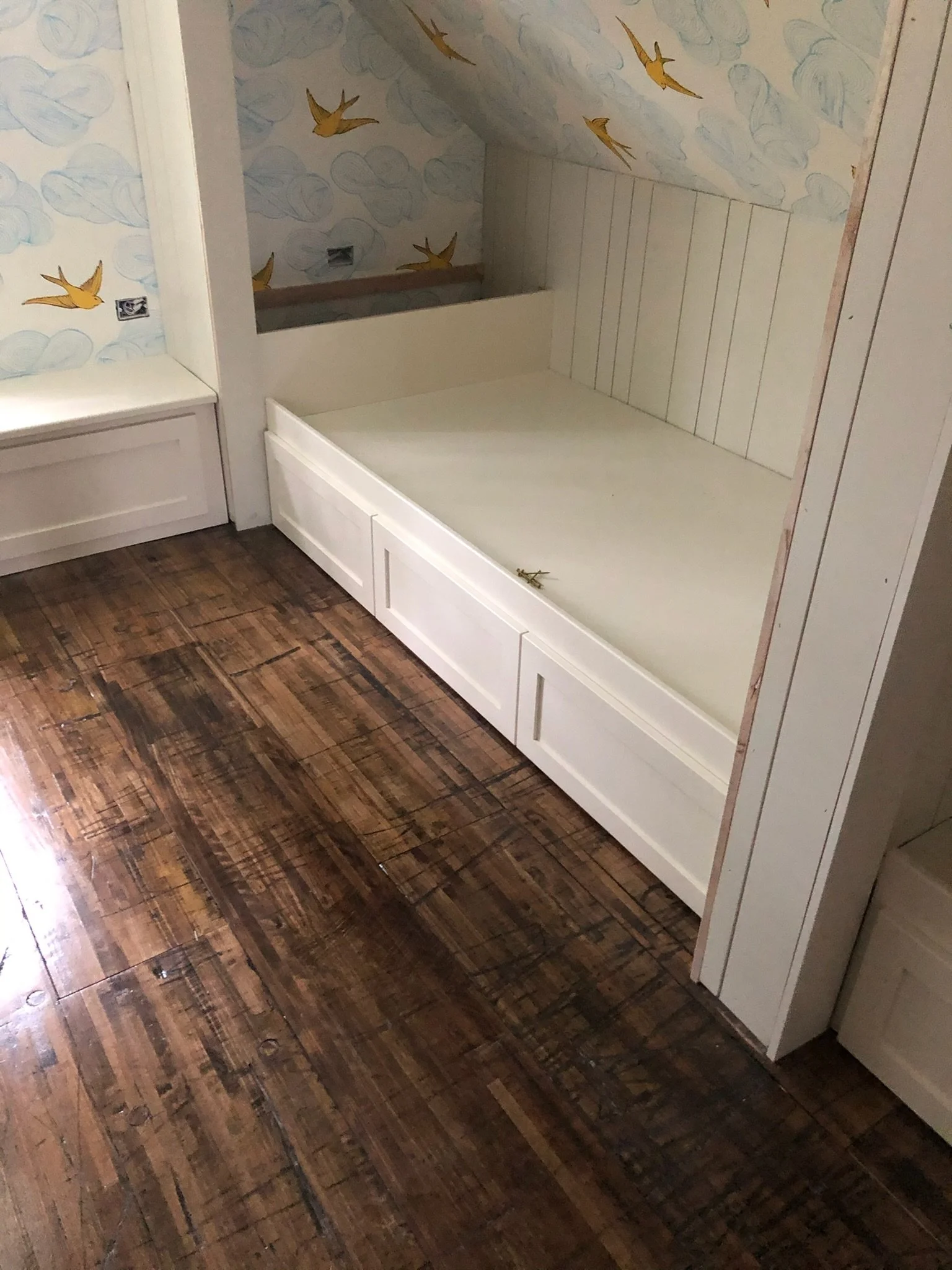 A built-in white bed with storage drawers underneath, positioned against a wall with cloud and bird wallpaper, in a room with wooden flooring.