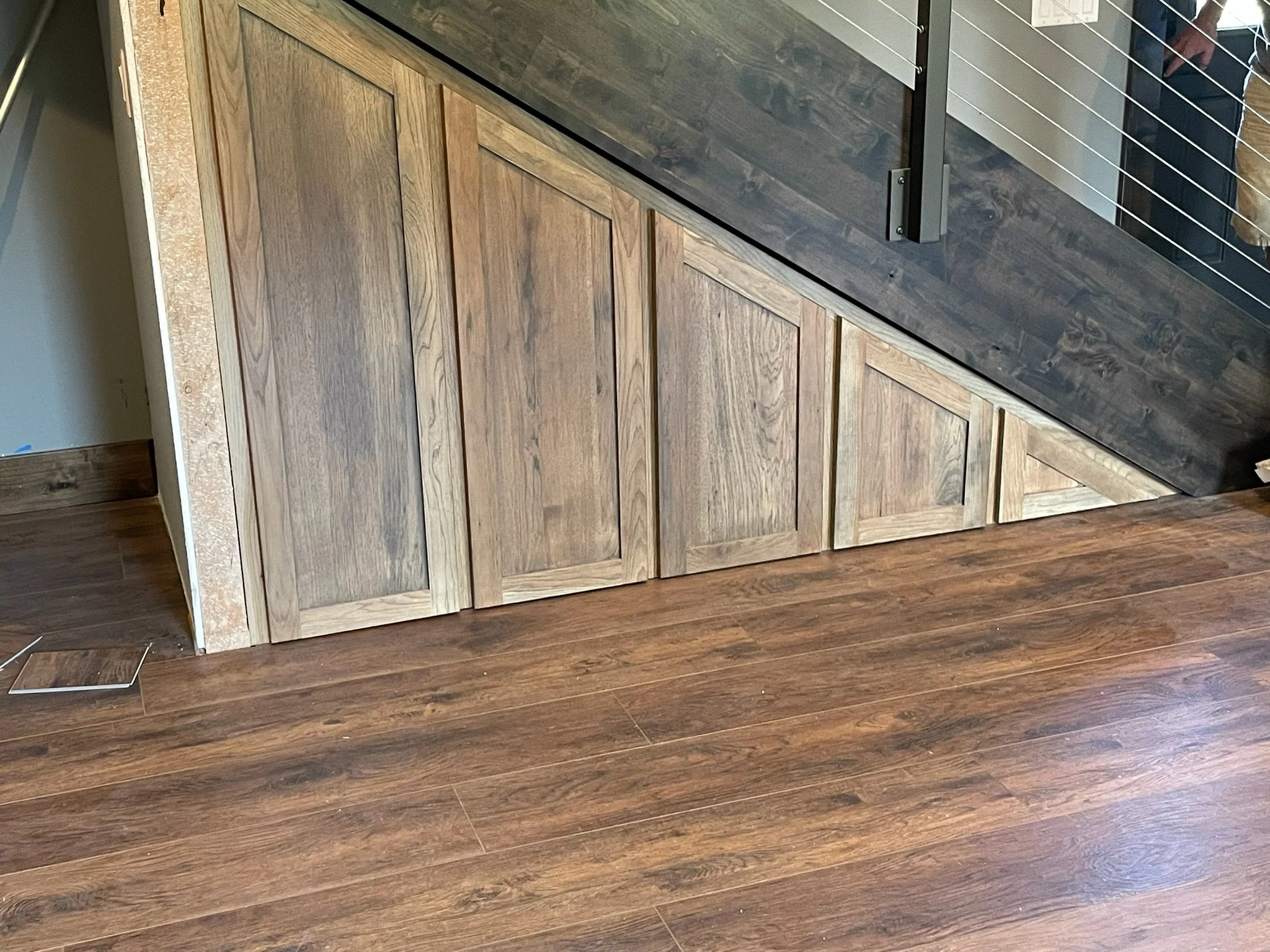 Close-up of hardwood floor and a wooden cabinet with a diagonal design, next to a section of darker wood flooring and a stair railing.