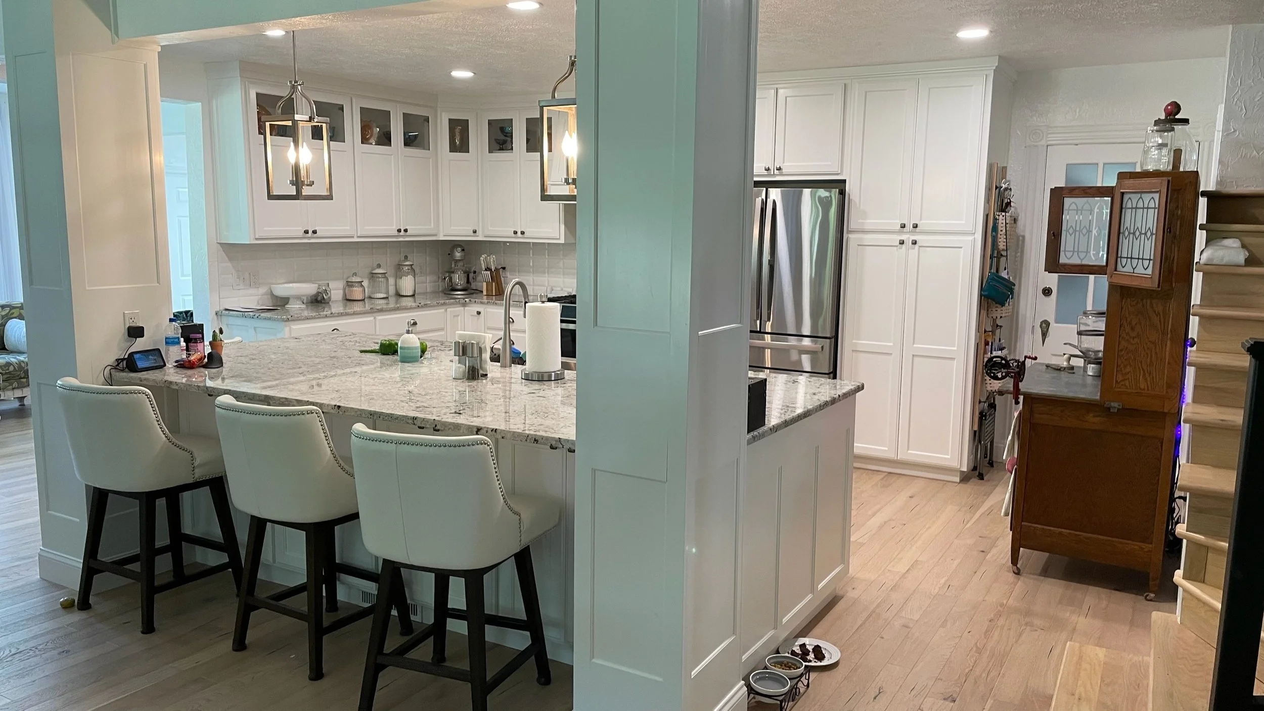 A modern kitchen with white cabinets, a marble island with three white bar stools, a stainless steel refrigerator, and various kitchen items on the counters. There are two light fixtures hanging above the island and a wooden cabinet near the staircas