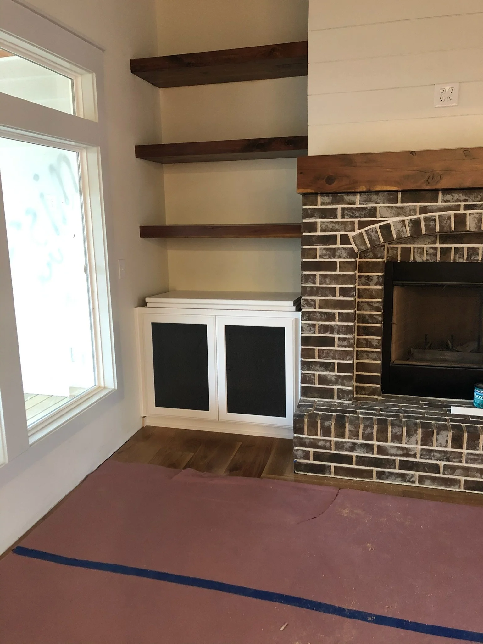 Corner of a living room featuring a brick fireplace, three wooden floating shelves on the wall, a white cabinet with black panels, a window, and a partially covered floor with a tarp.