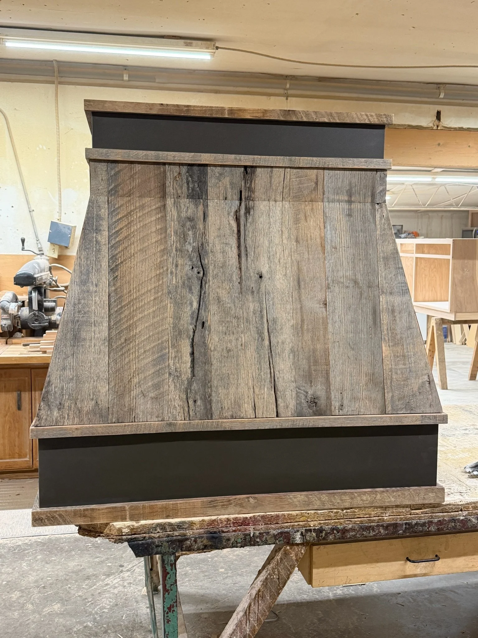 A wooden cabinet or furniture piece in a workshop with finished wood surfaces on top and bottom and a black painted section in the middle, with visible wood grain and cracks.