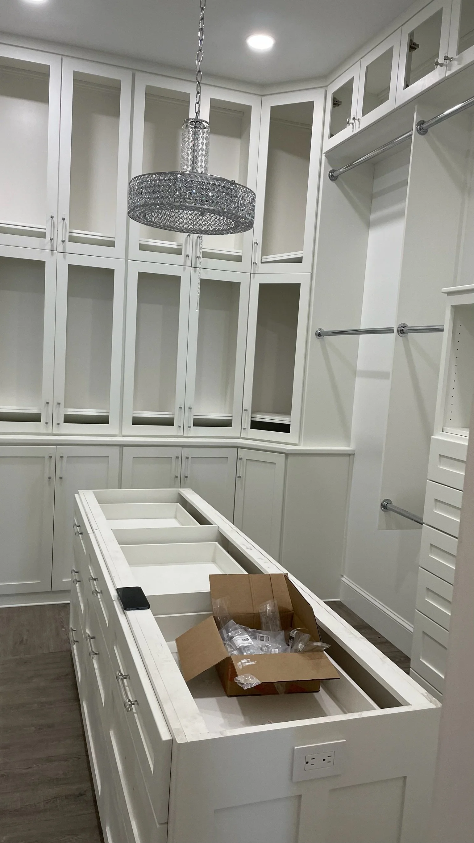 White walk-in closet with empty cabinetry, a chandelier hanging from the ceiling, and a large, empty island in the center with a cardboard box and plastic packaging inside.