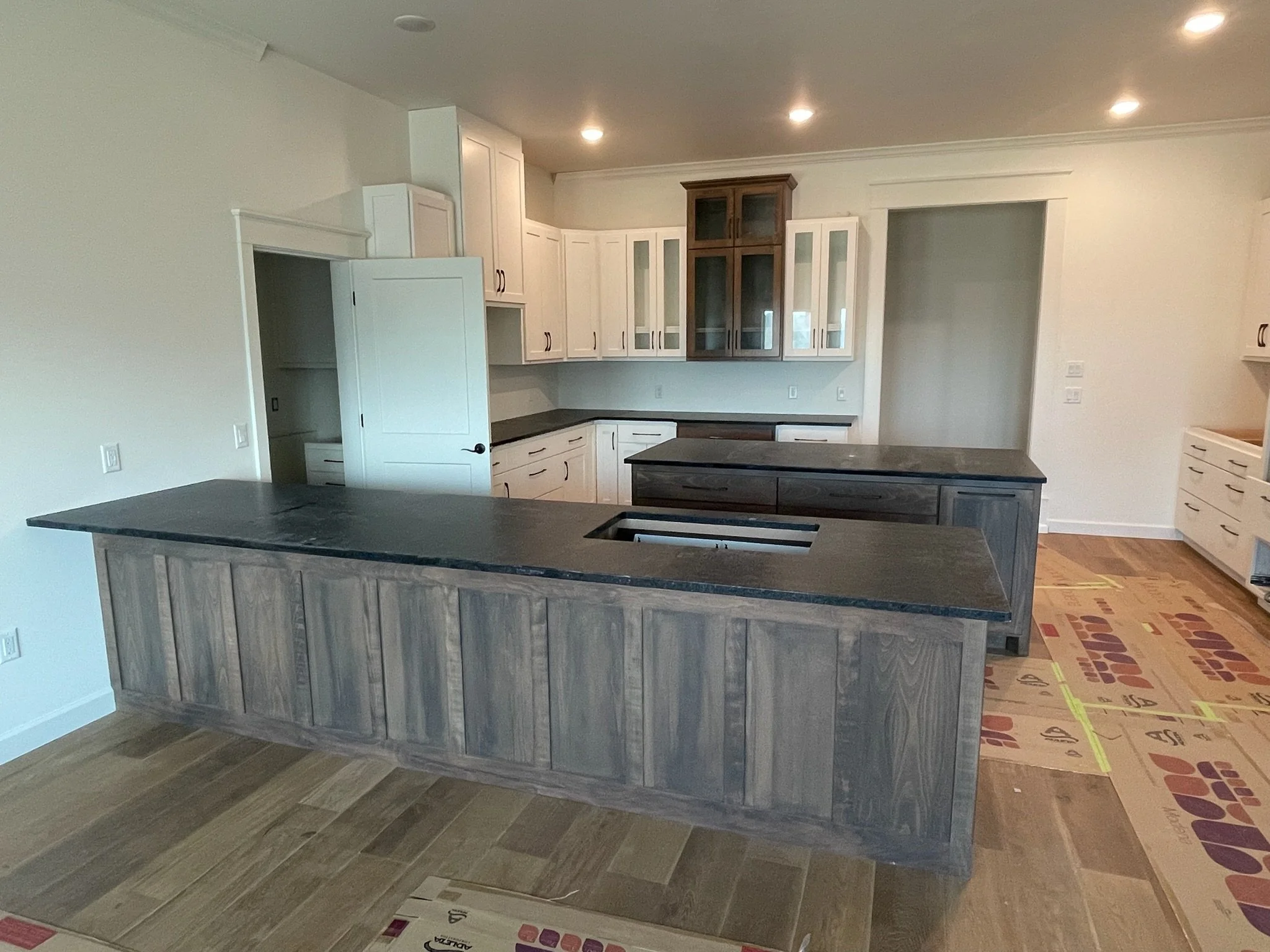 Empty kitchen with black countertops, white upper cabinets, dark lower cabinet island, and hardwood flooring.