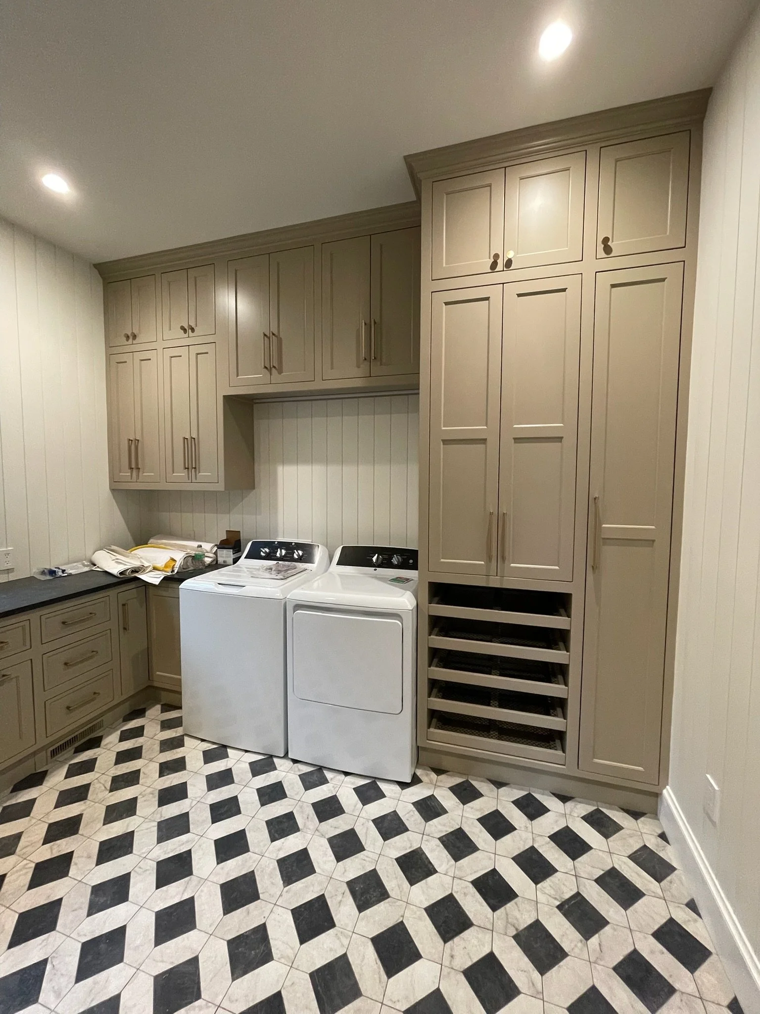 Laundry room with beige cabinets, a washing machine, and dryer, patterned black and white floor tiles, and some papers on the counter.