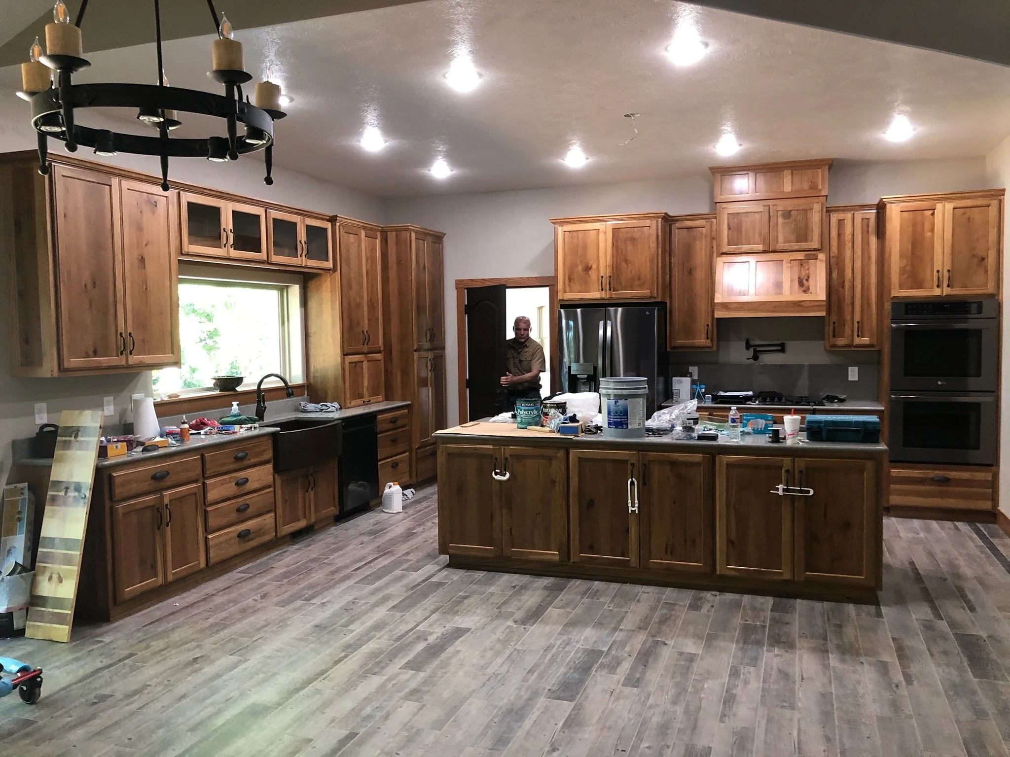 A spacious kitchen with wooden cabinets, a large island, stainless steel appliances, and a man standing near the doorway. The kitchen is under renovation, with tools and materials on the countertops.