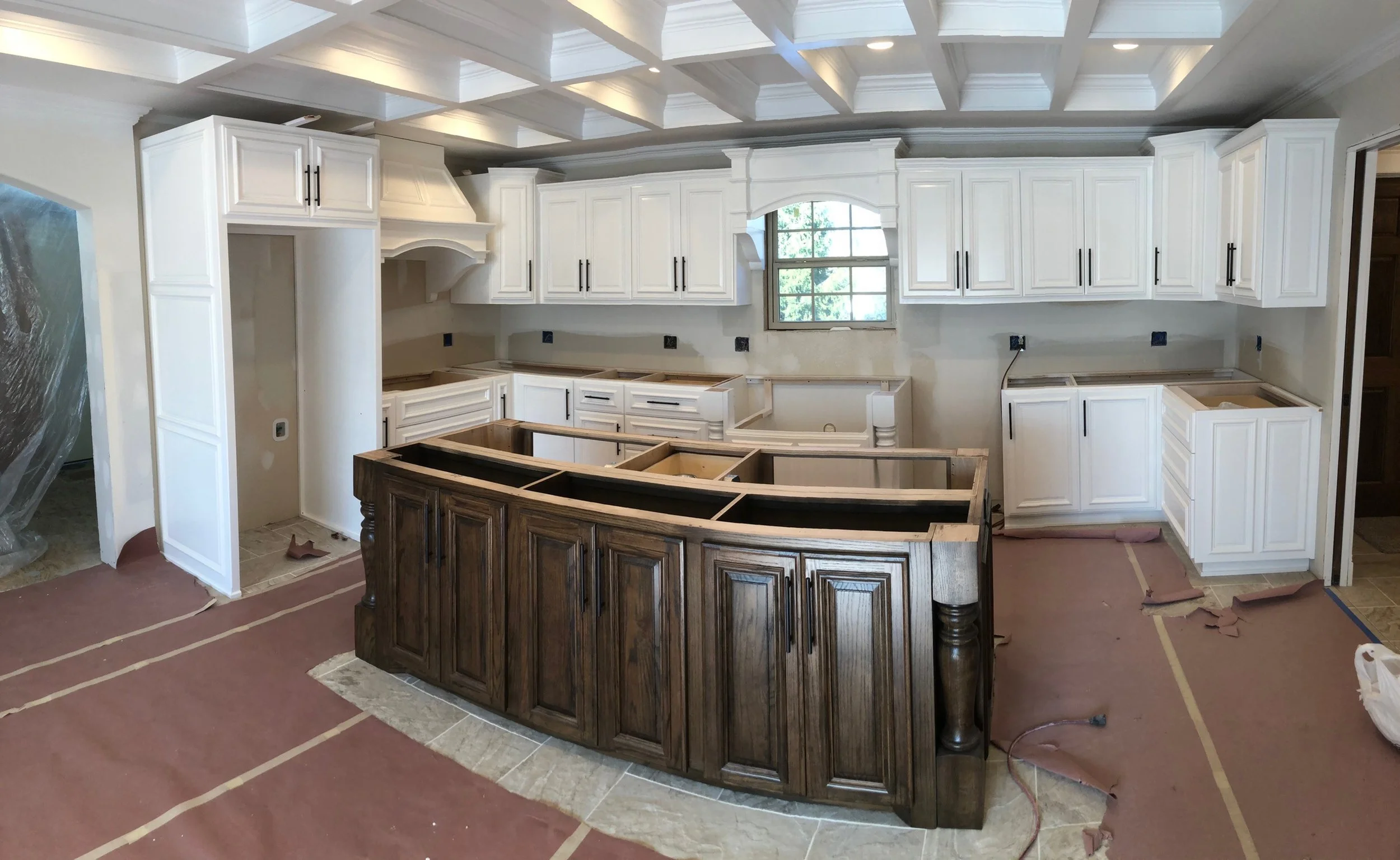 Kitchen under construction with white upper cabinets, a large window, and a dark wood curved island in progress.