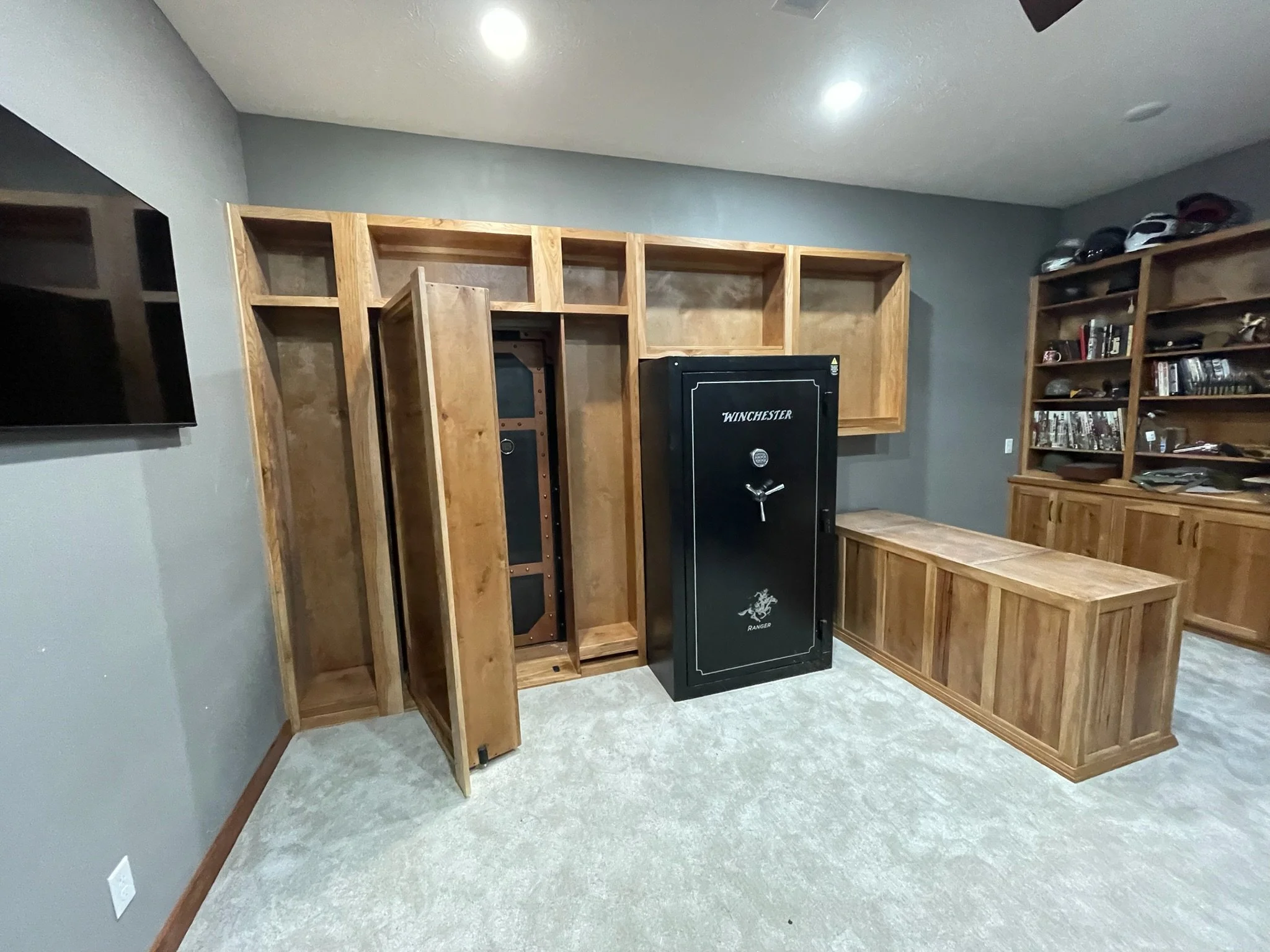 A room with gray walls and beige carpet, featuring wooden shelves and cabinets, a black Winchester safe in the center, and a flat-screen TV mounted on the wall to the left.