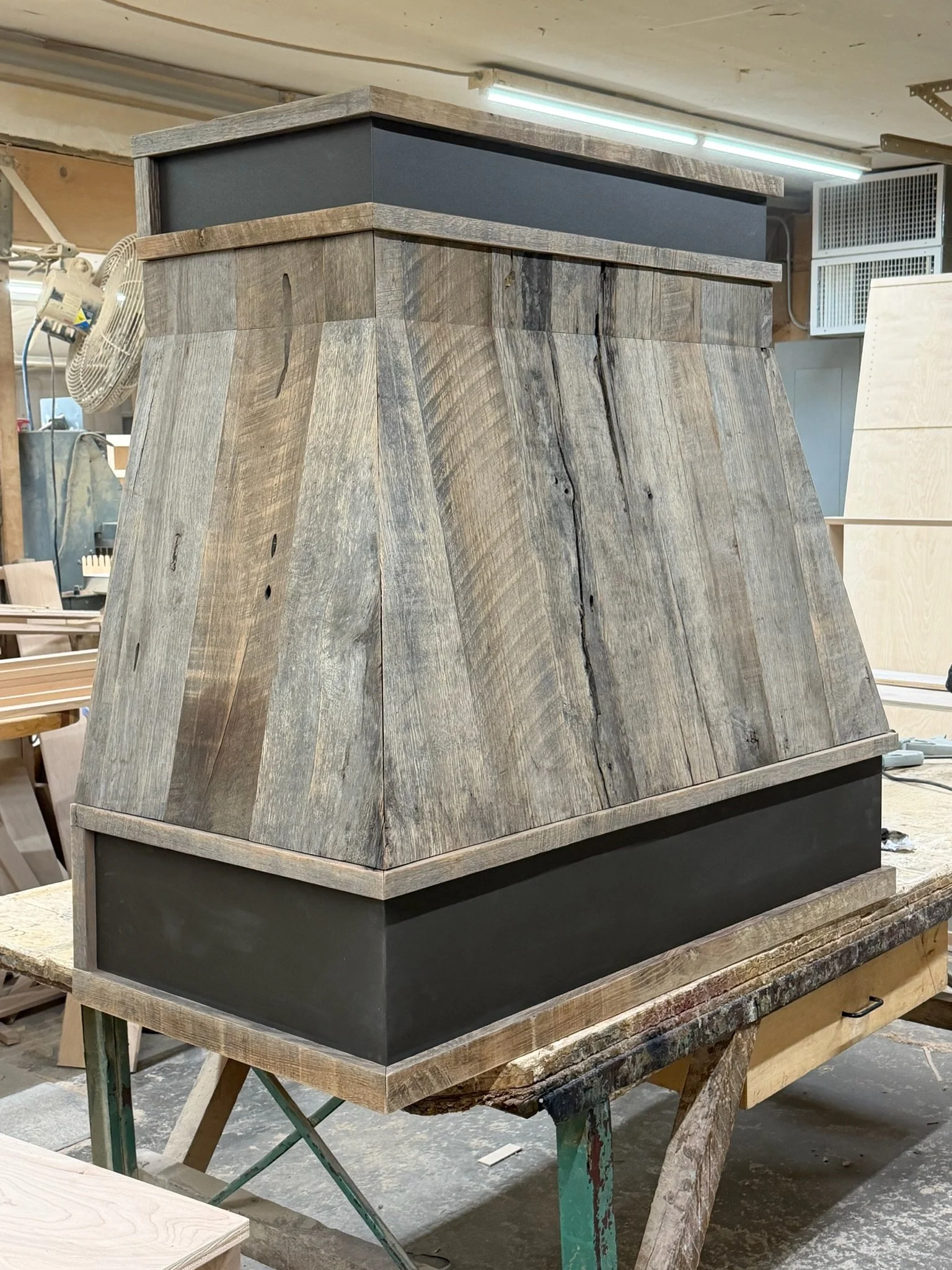 A wooden kitchen range hood with a black trim, situated on a workbench in a woodworking shop.