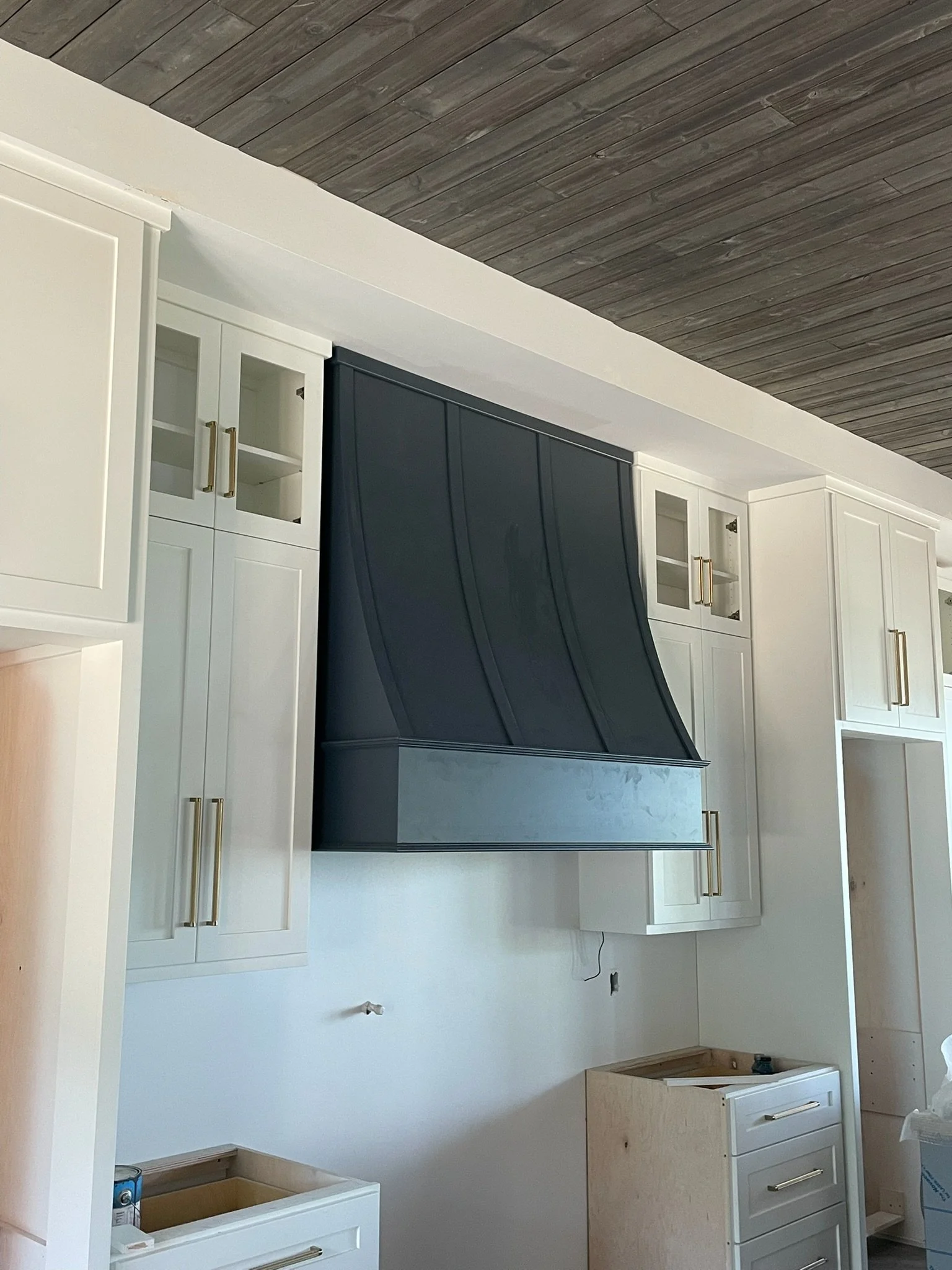 Kitchen under construction with white cabinets, a black range hood, and wooden ceiling.