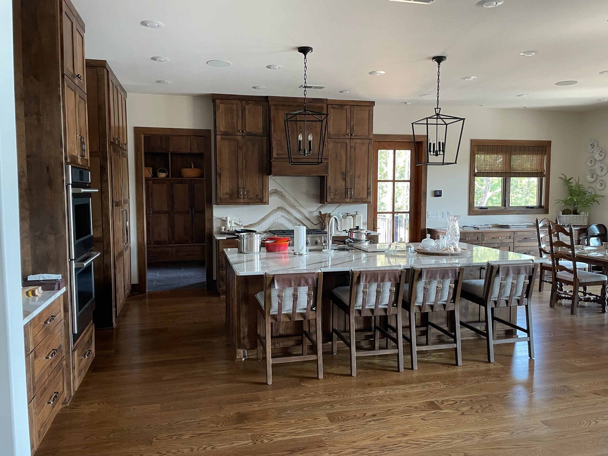 A spacious kitchen with wooden cabinets, a large island with bar stools, a double oven, modern pendant lights, and a dining area with a table and chairs. There are windows allowing natural light, and the floor is hardwood.