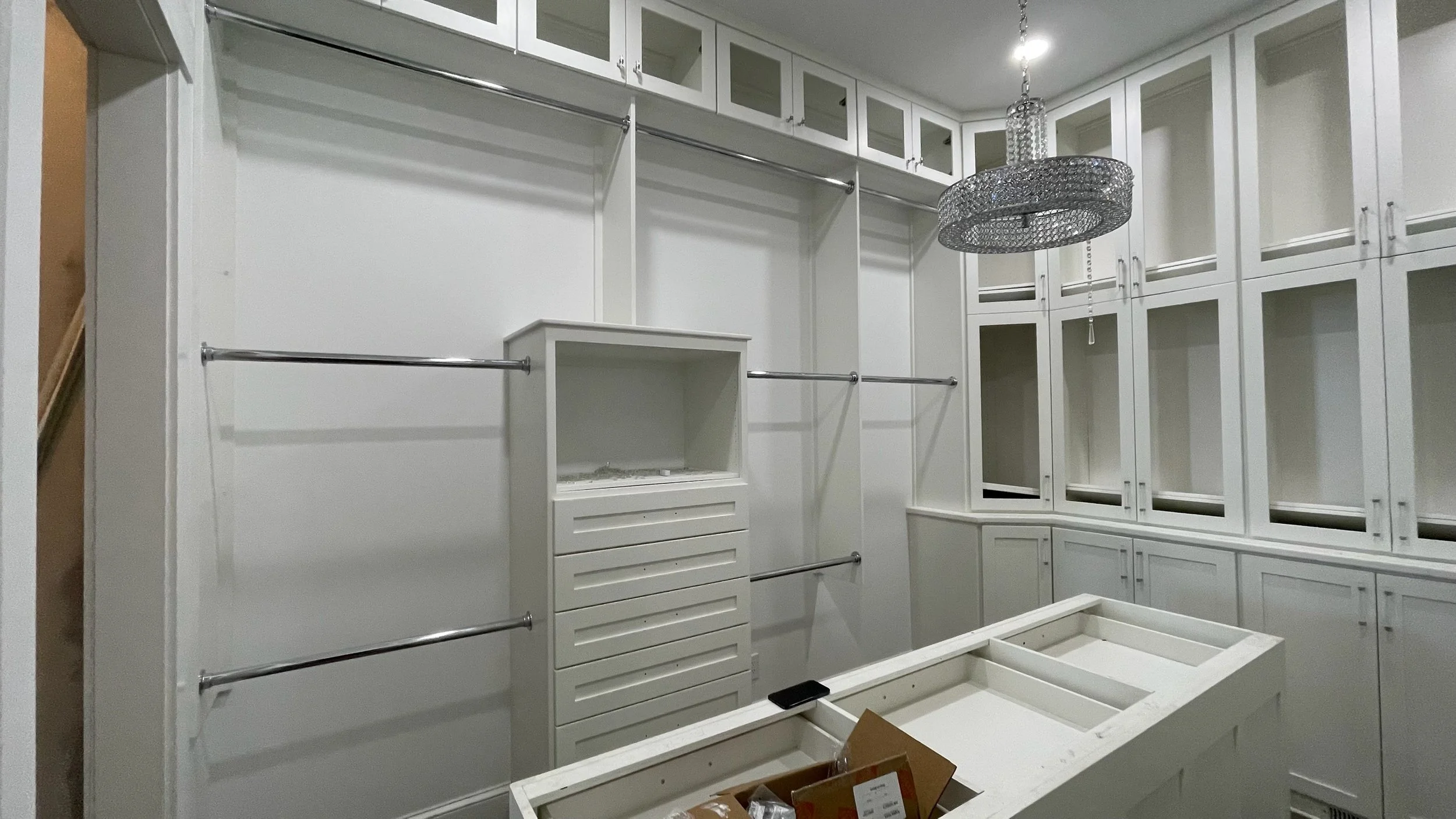 A white walk-in closet with built-in cabinets, shelves, and hanging rods; a chandelier hanging from the ceiling; an island with open compartments in the center; and an unfinished cabinet or drawer on the floor.