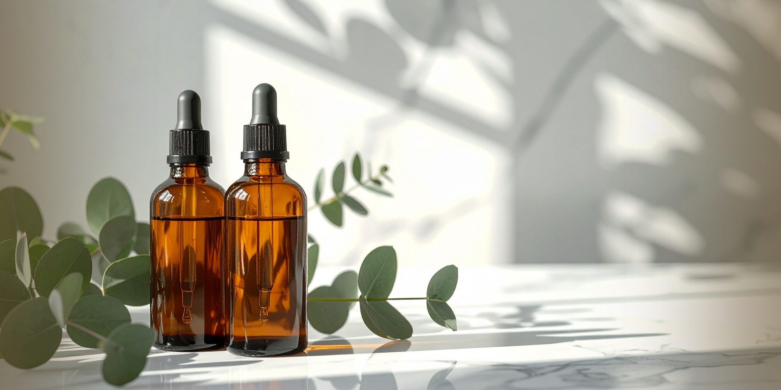 Two amber glass dropper bottles with black caps and droppers, set among green eucalyptus leaves with shadows cast on a white surface in natural light.