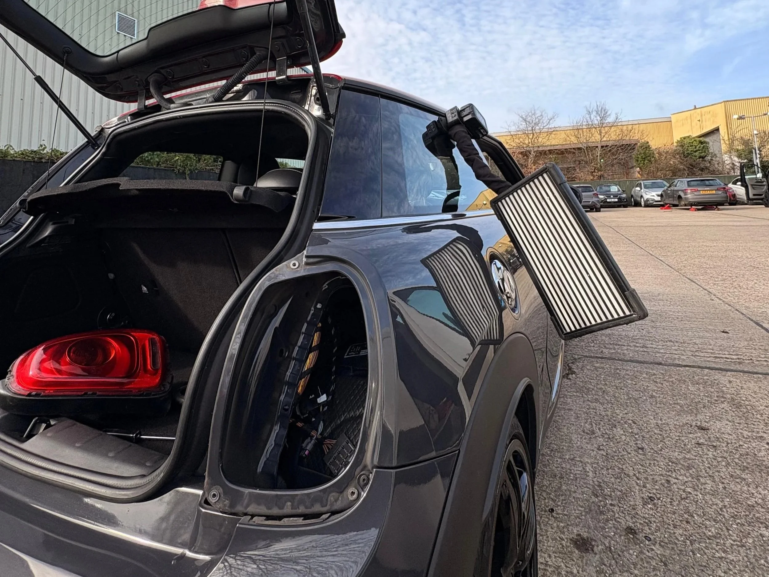 An electric car with its rear hatch open and a large light panel attached to the side, parked in a lot with other vehicles and a yellow building in the background.