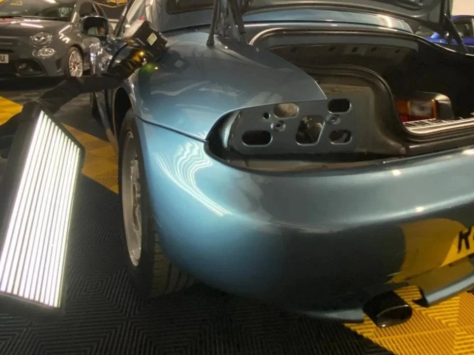 A blue sports car with its rear removed, exposing the empty taillight housing and internal components. The car is parked indoors with other vehicles visible in the background.