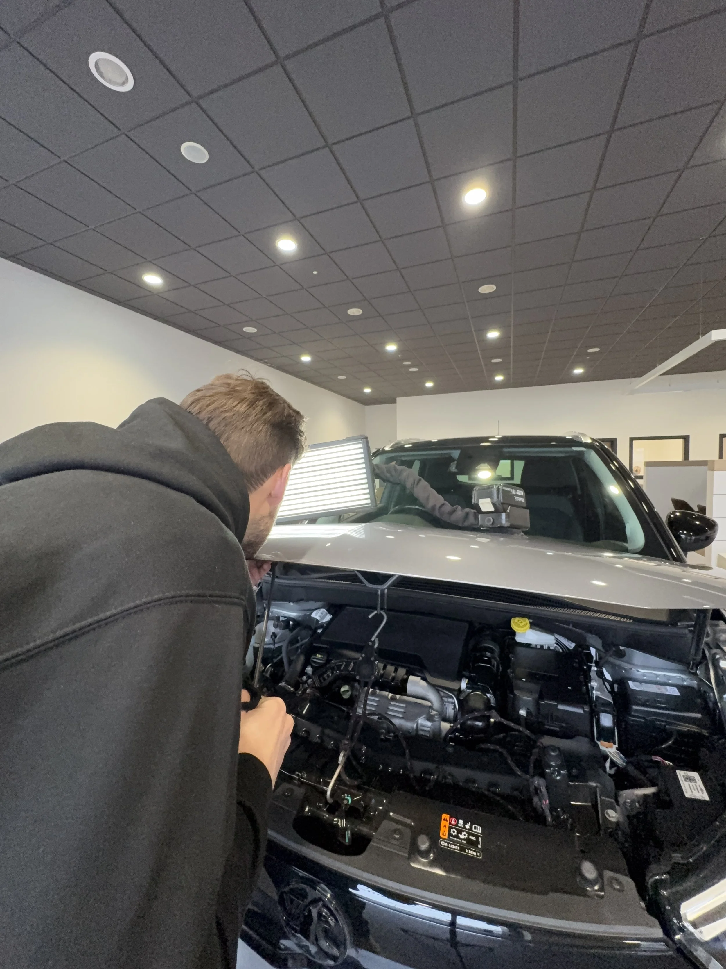 A man inspecting the engine of a car inside a dealership or garage, with a worker in the background.